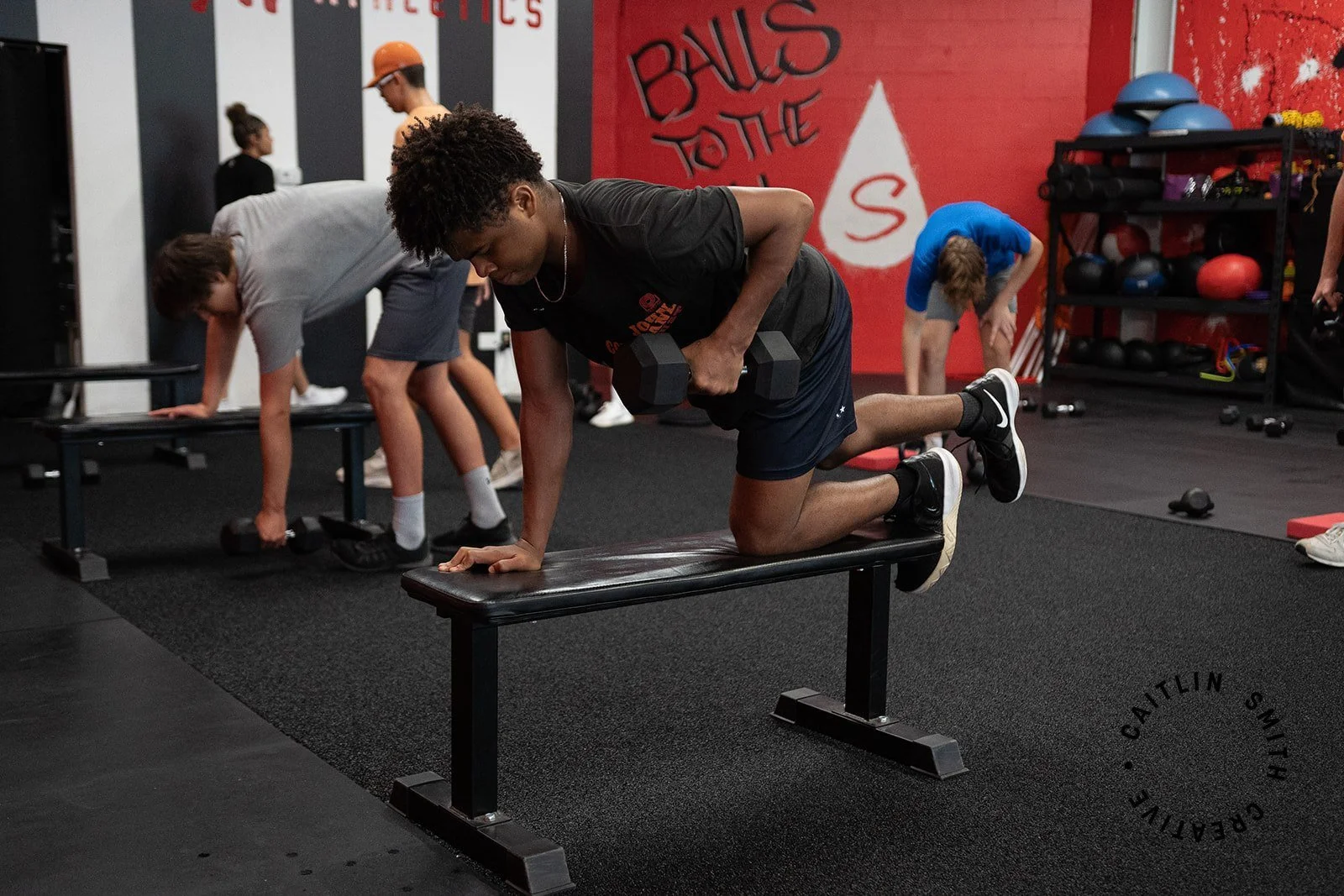 A young man doing a one-arm push-up with his knee on a bench, holding a dumbbell in his other hand at a gym. Other people are working out in the background.