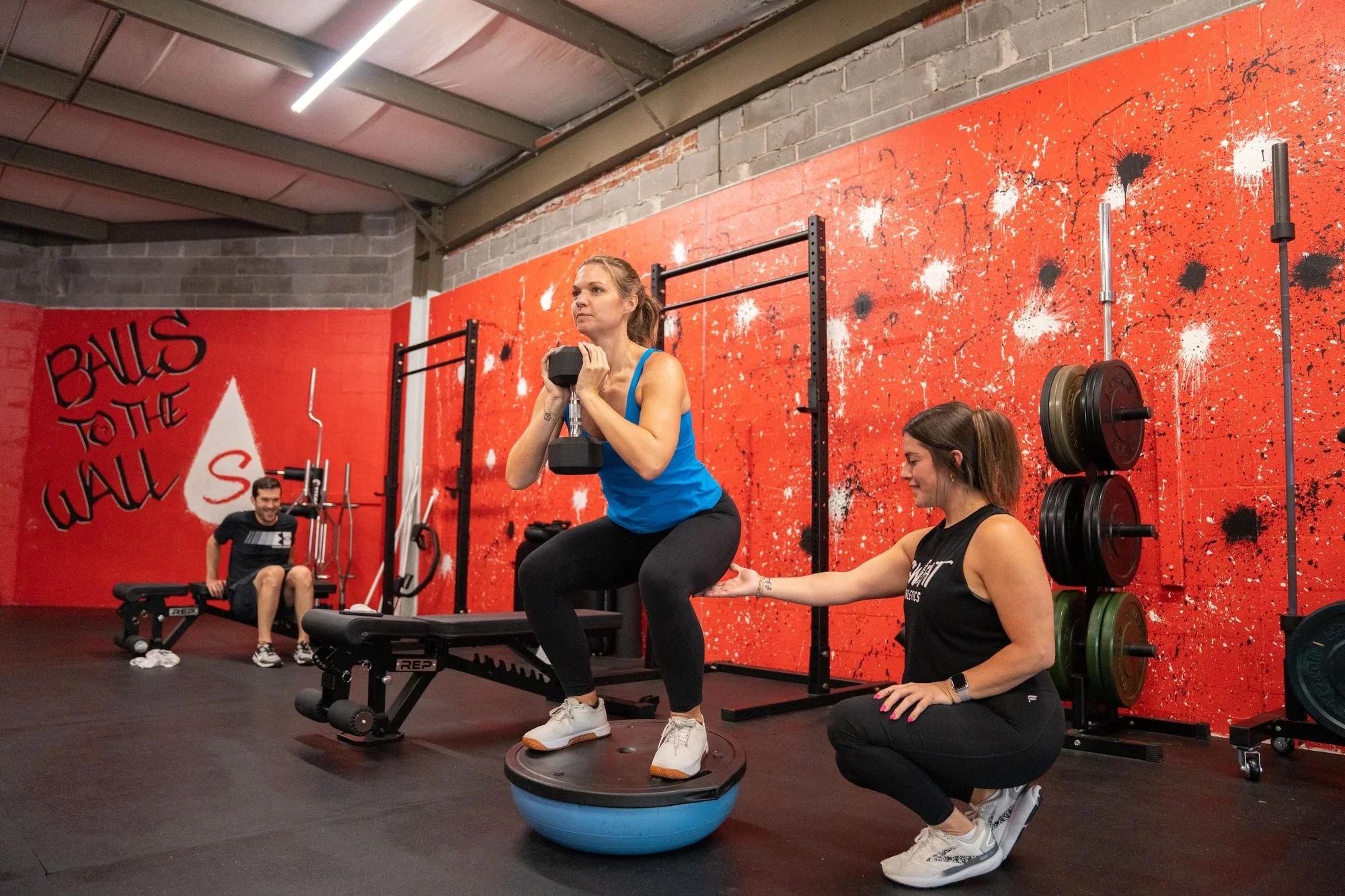 Women in a gym exercise class performing squats on a purple exercise ball with a trainer assisting. Other participants sit on benches and weights, with a red splash wall background.
