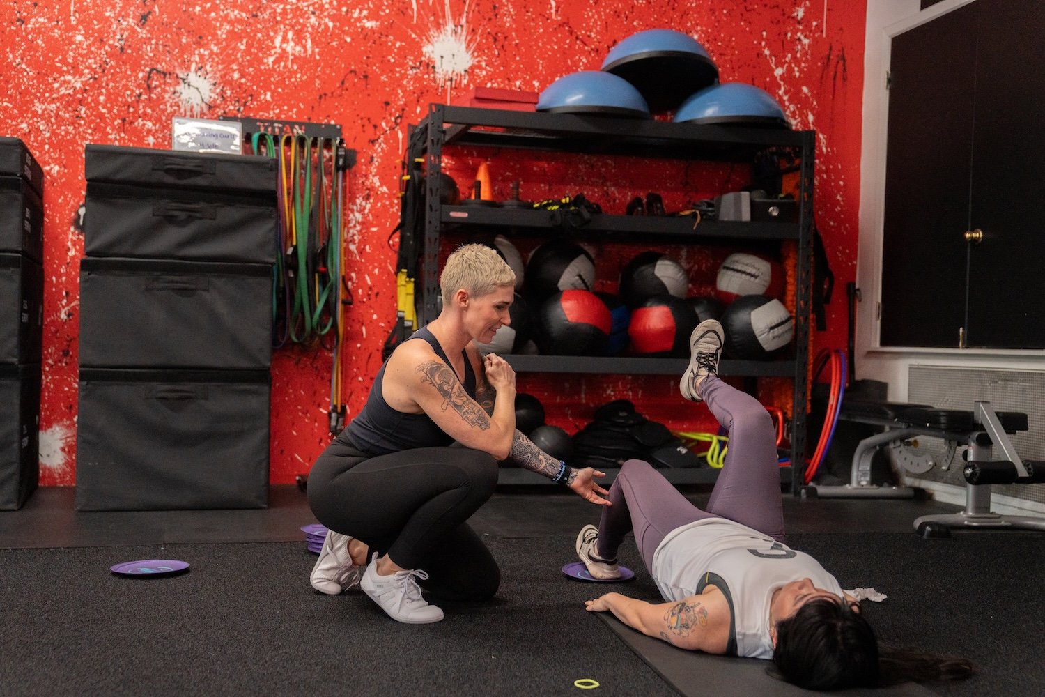 A fitness trainer helping a woman stretch on gym floor, with workout equipment like medicine balls, resistance bands, and balance balls in the background.