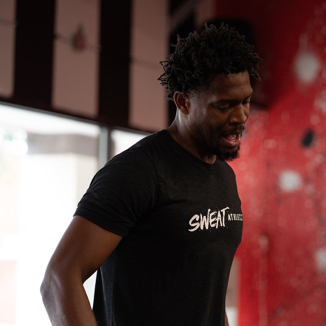 A man with curly hair and a beard, wearing a black t-shirt that says "SWEAT ATHLETICS," appears to be working out or exercising in a gym or fitness center.