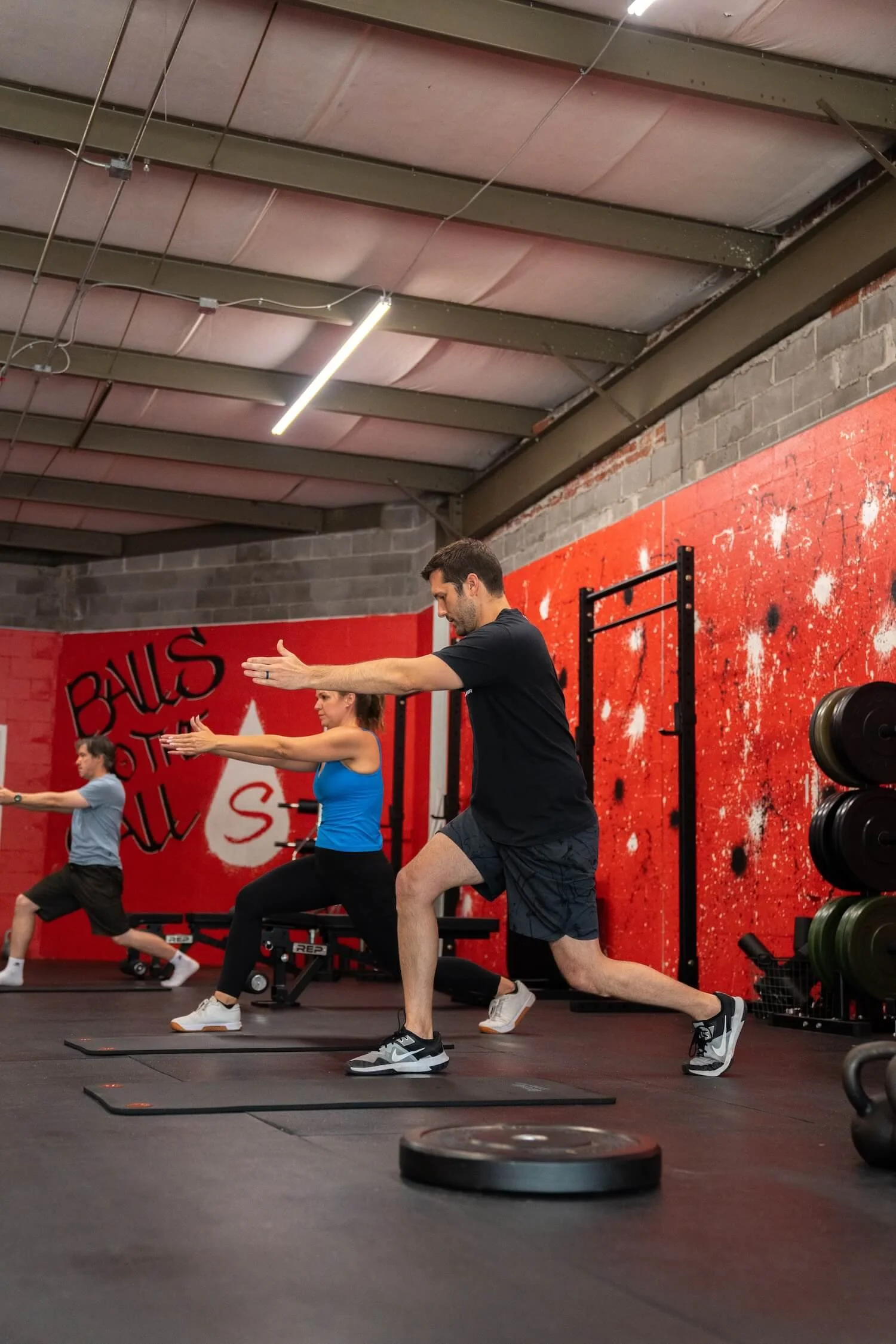 People working out in a gym, performing lunges on black exercise mats in front of a red and gray exercise wall.