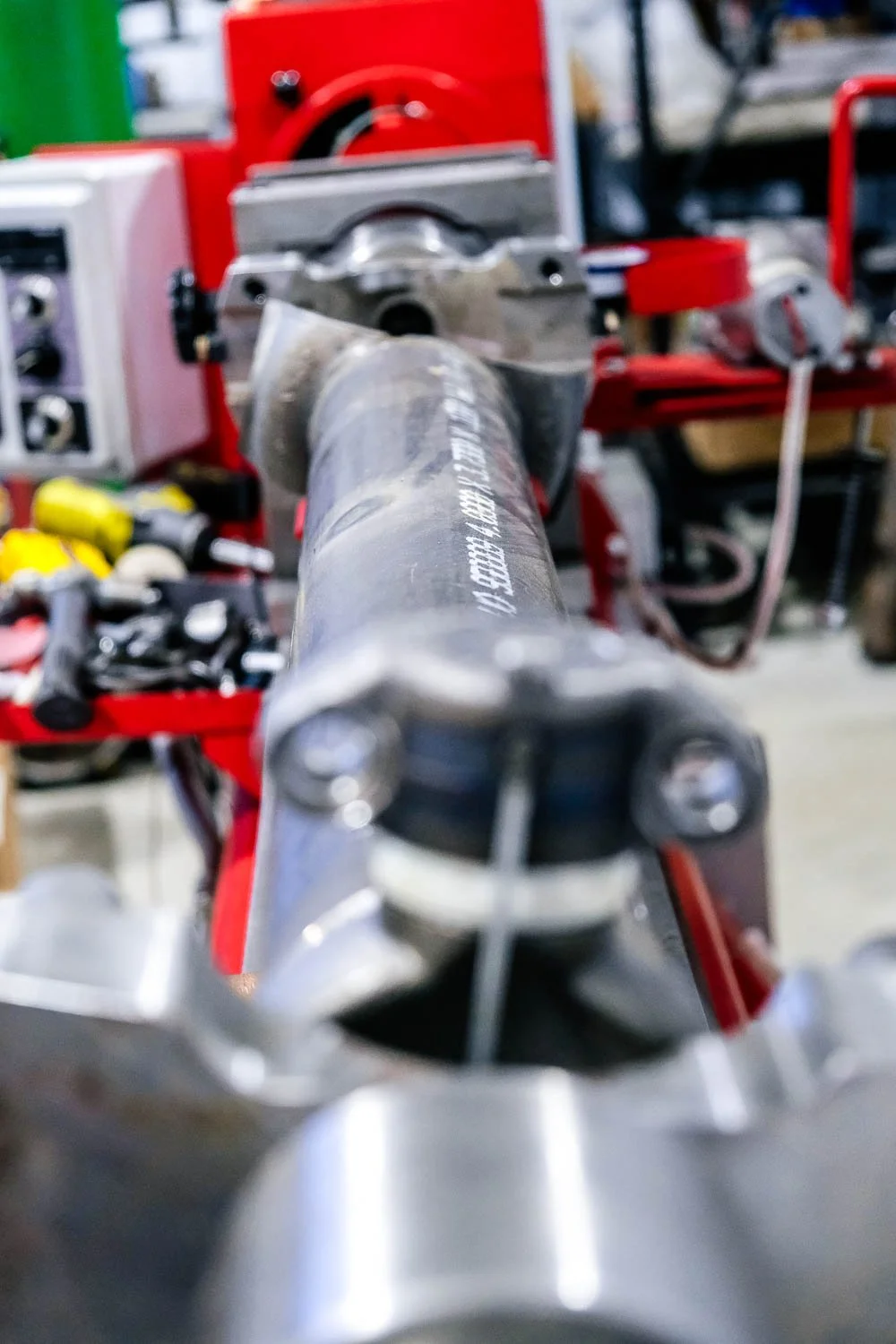 Close-up view of a metal tool or part positioned on a workbench, with various workshop tools and equipment in the background.
