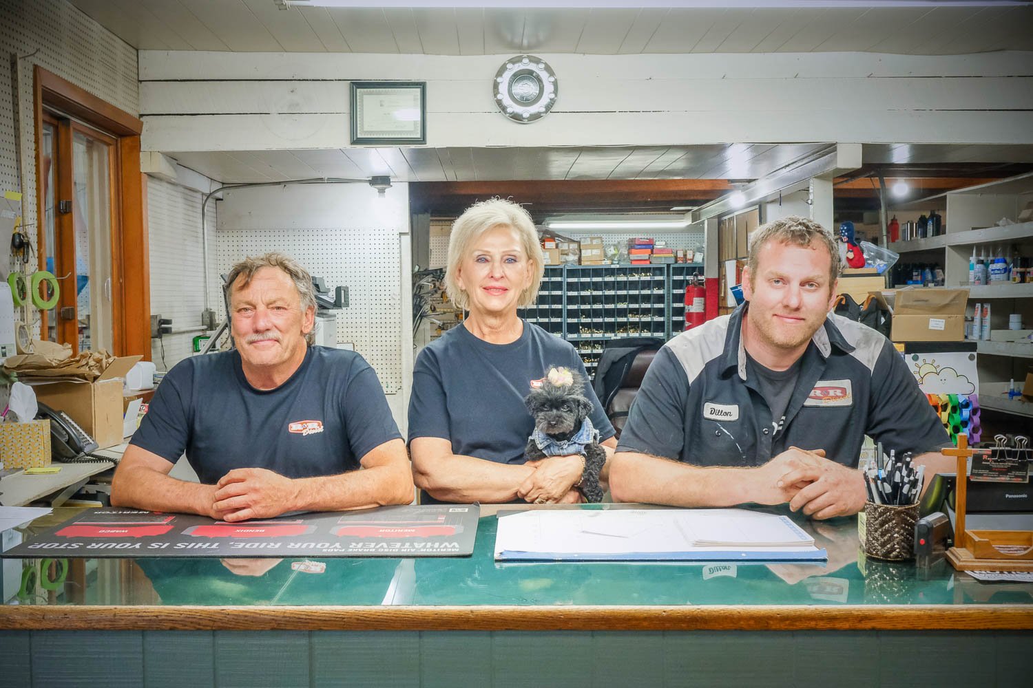 Three people and a dog sitting at a counter inside a store or workshop. Two men and a woman, with the woman holding a small dog wearing a bow. Shelves and various supplies are visible in the background.