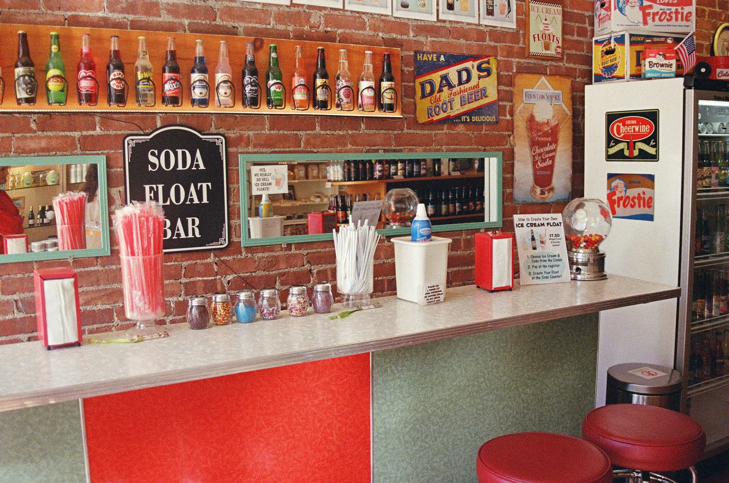 A counter at an ice cream shop with soda bottles on a wall shelf, colorful jars of sprinkles, a sign advertising a soda float bar, and various ice cream-related posters and signs.