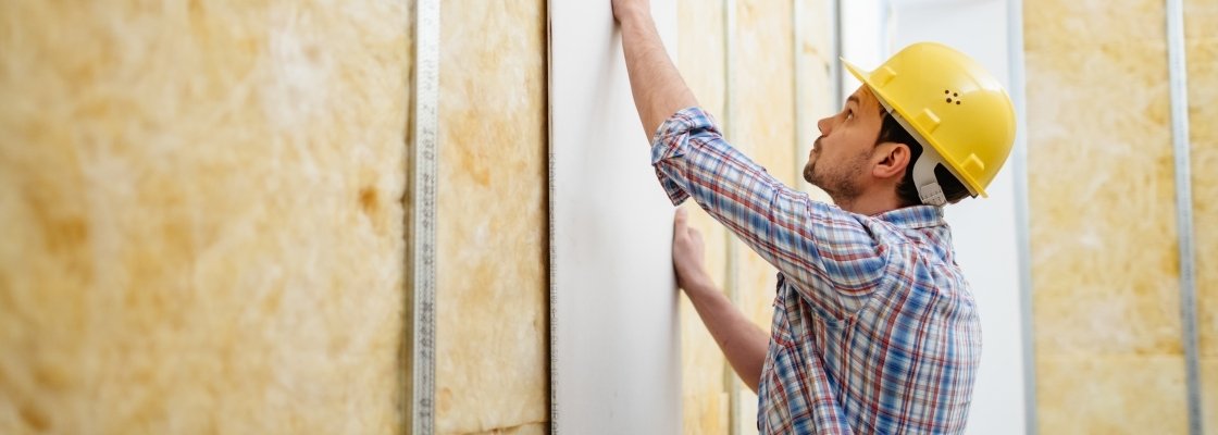 A construction worker wearing a yellow hard hat and plaid shirt installing drywall on a wall.