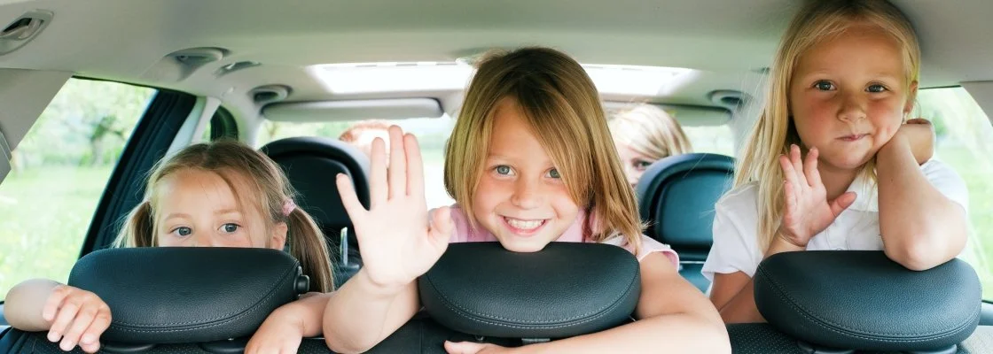 Four children looking out from the backseat of a car, with one girl waving at the camera.