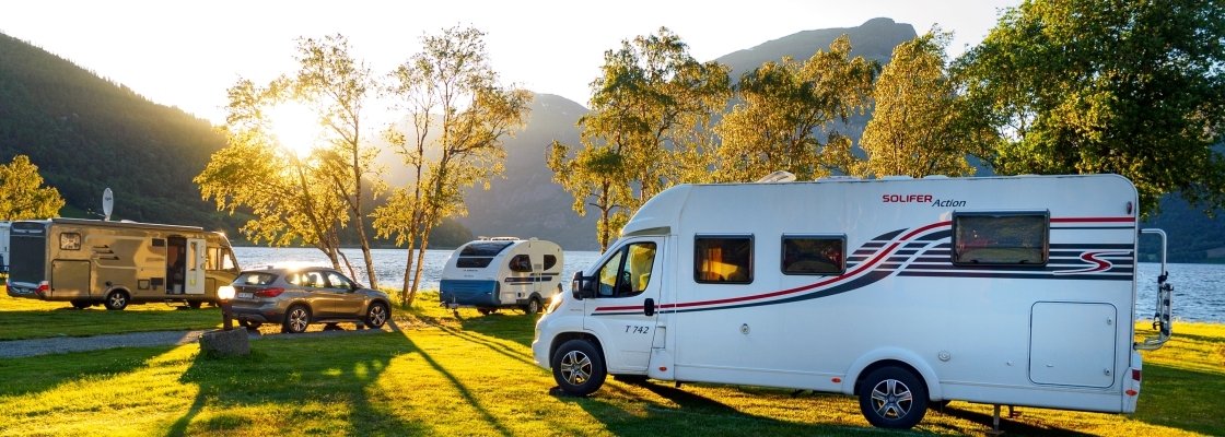 Campground by a lake with three RVs parked on grass, trees, mountains, and a sunset in the background.