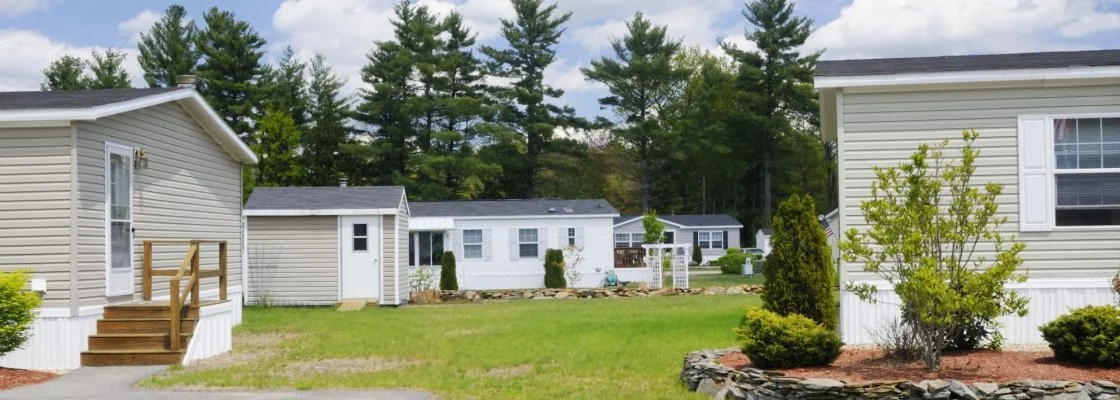 A row of mobile homes with front yards and a grassy field, surrounded by trees.