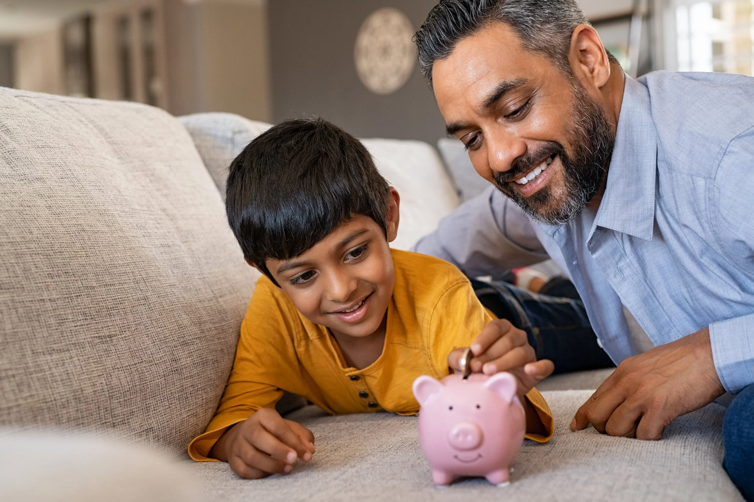 A smiling man with gray hair and beard, and a young boy with dark hair, are sitting on a beige couch, putting money into a pink piggy bank on the floor in front of them.
