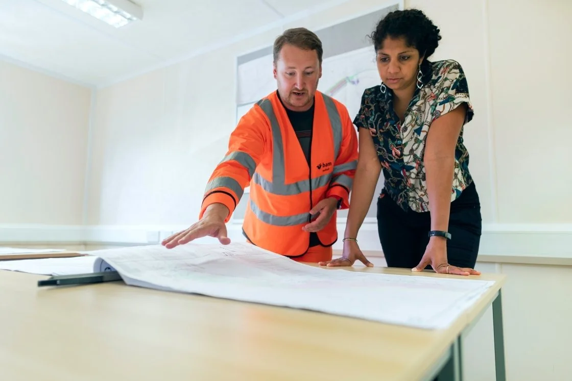 Two women look at blueprints on a table in an office, one wearing an orange safety vest.