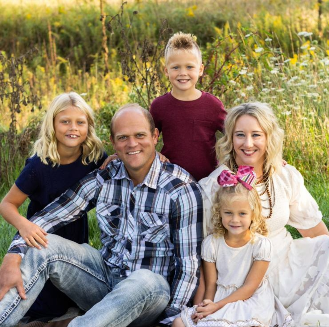 A family of six sitting outdoors in a grassy meadow with tall plants and trees in the background, smiling for a photo.