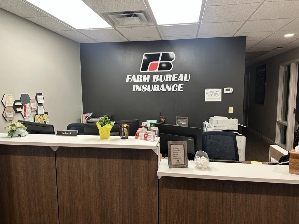 Office reception area for Farm Bureau Insurance with a dark gray wall displaying the company logo and name, a white reception desk with various office supplies, plants, and decorations, and a printer to the side.