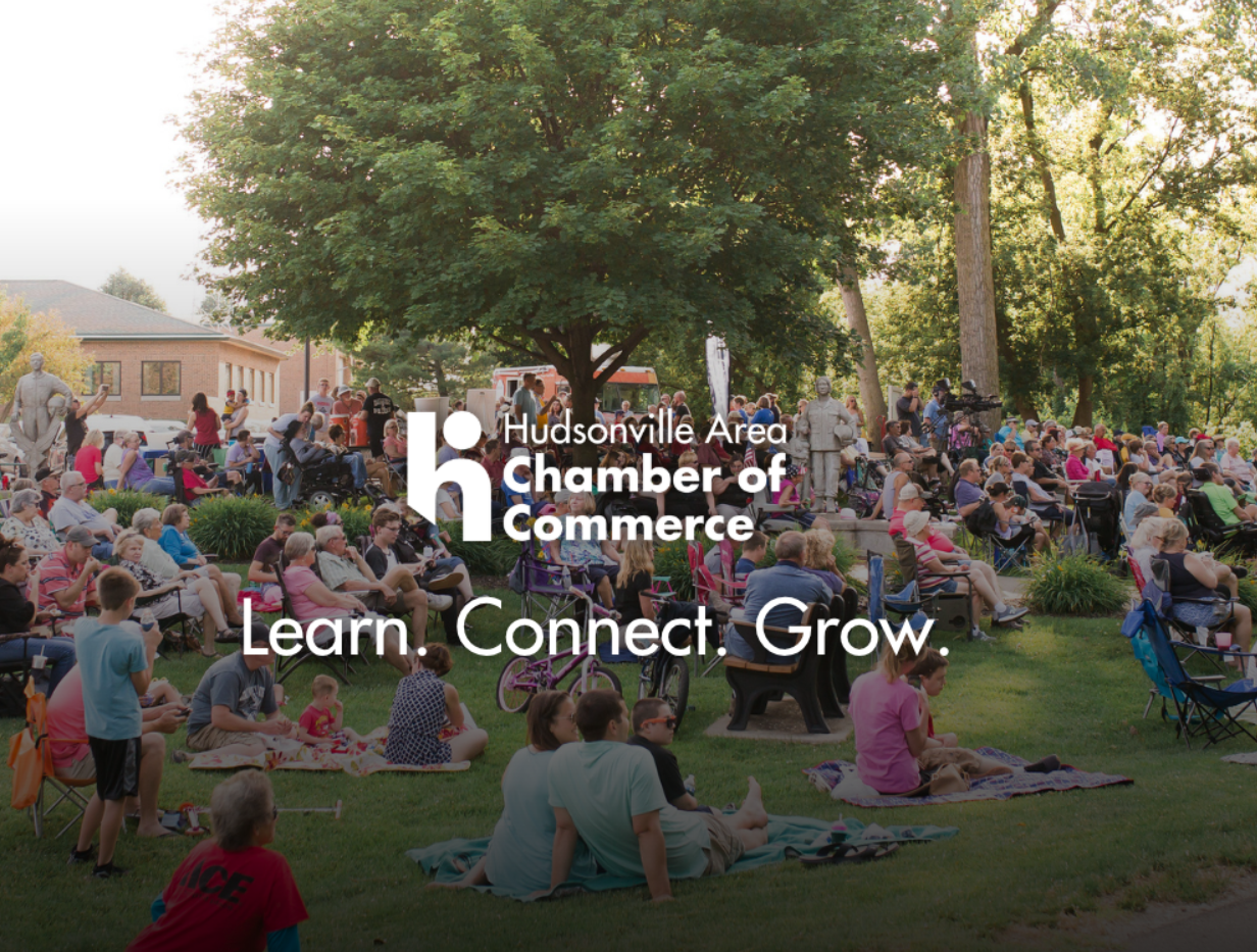 A large crowd gathered outdoors on a lawn under trees during daytime, attending an event organized by the Hudsonville Area Chamber of Commerce. People are seated on chairs and blankets, with some standing. The scene includes a statue, a fire truck in the background, and a mix of adults and children enjoying the event.