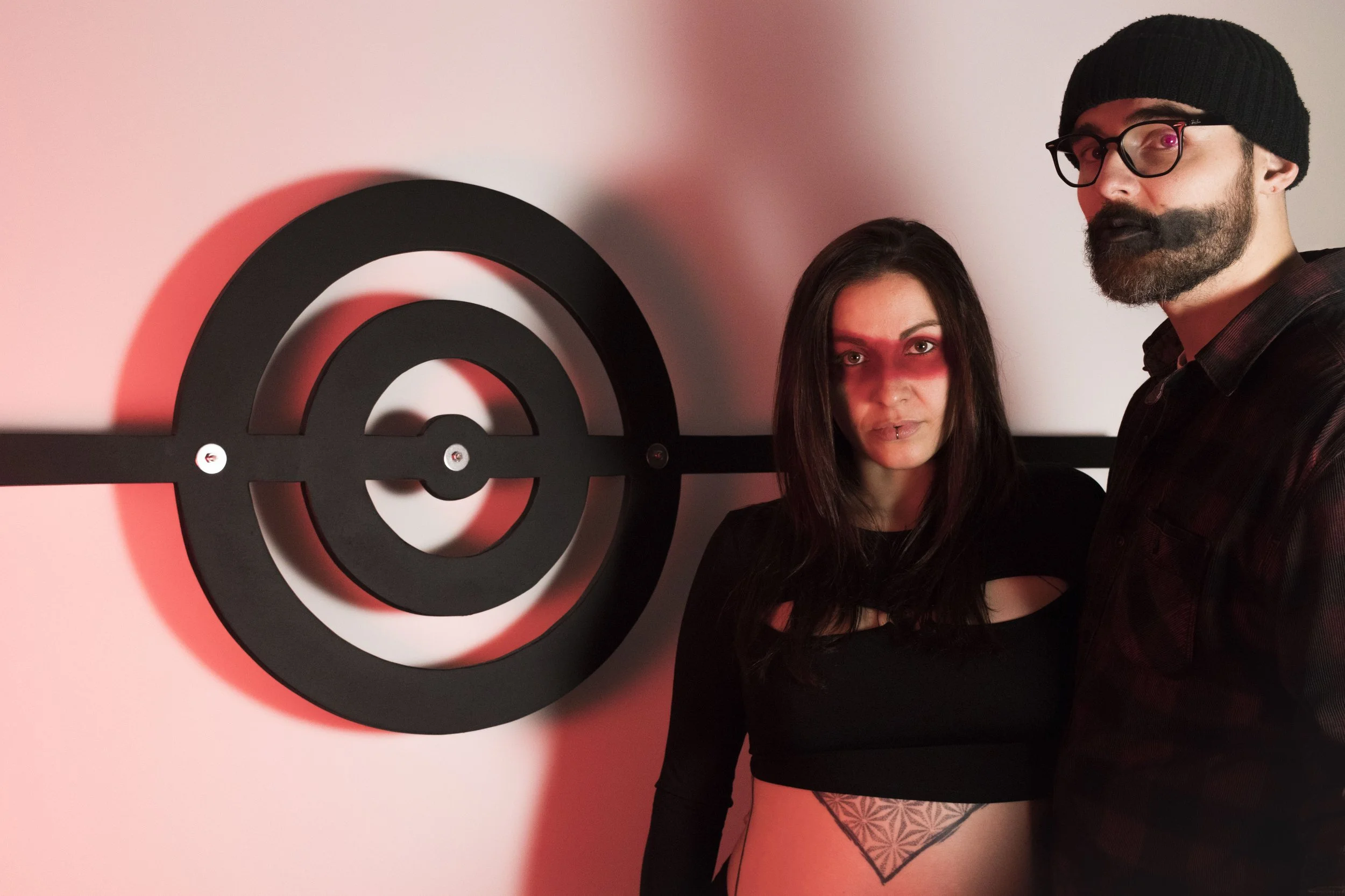 Olivier et caroline poinsignon à coté du logo Oriel dans leur atelier avec une lumière rouge pour constituer les ombres. Maquillage noir pour Olivier et rouge pour Caroline.
