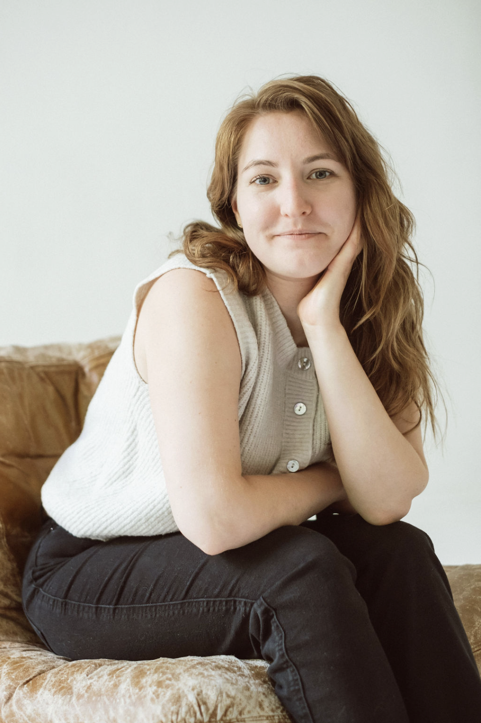 A young woman with wavy red hair, wearing a sleeveless light-colored knit top, sits on a brown velvet couch with a neutral expression, resting her chin on her hand, against a plain white wall.