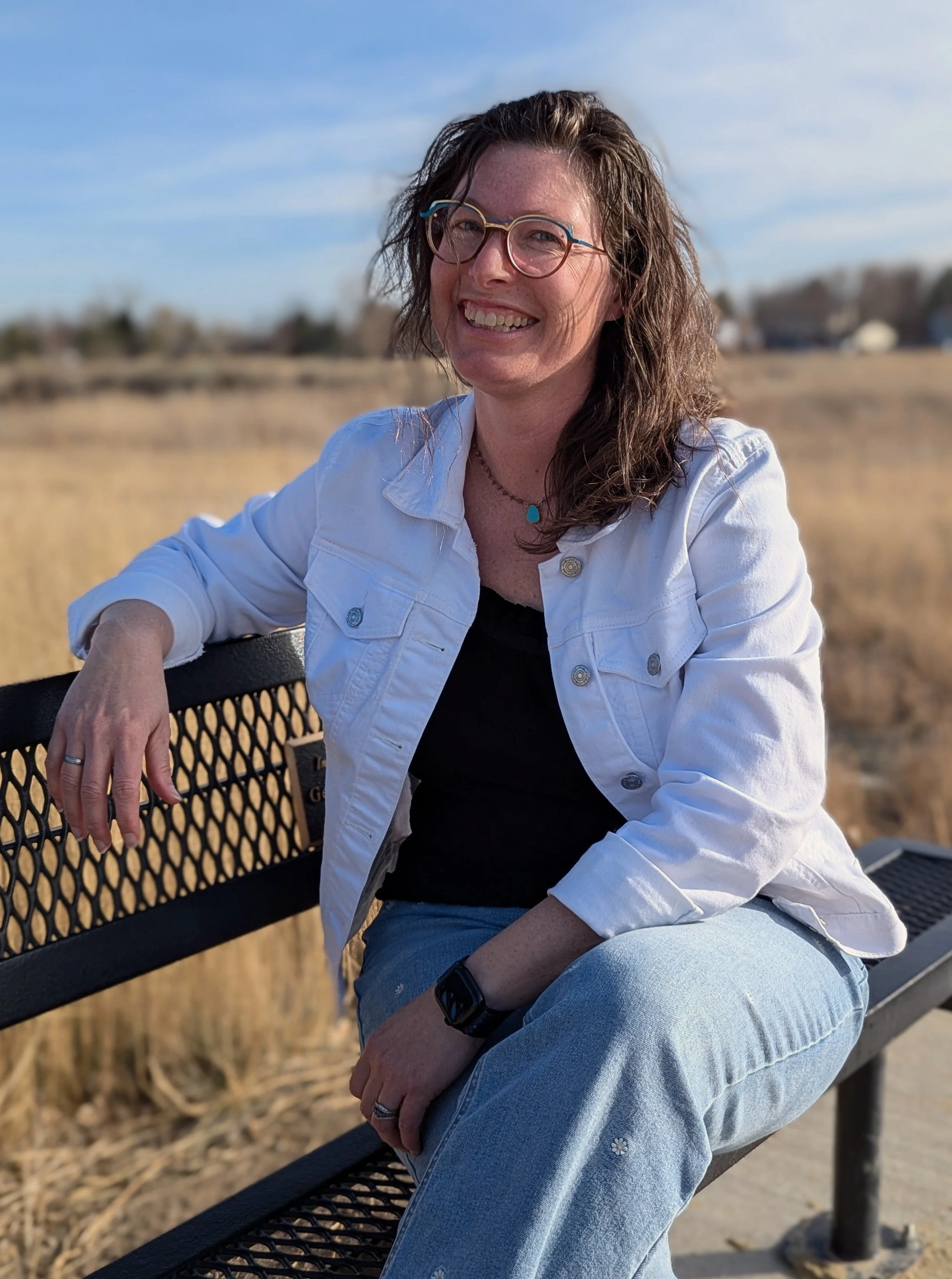 The author with curly brown hair and glasses smiling while sitting on a park bench with a field and houses in the background on a sunny day.