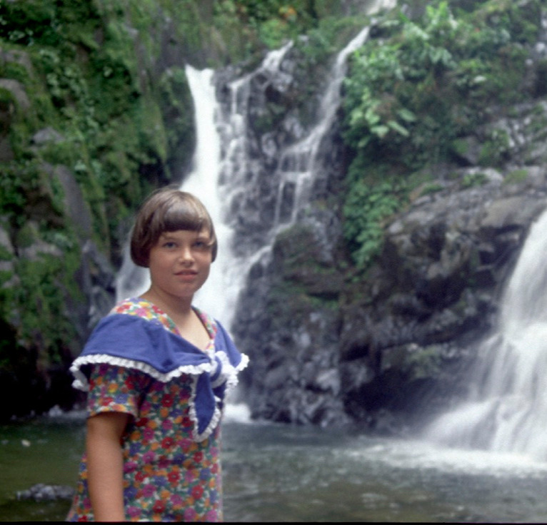 Young girl with short brown hair standing in front of a waterfall flowing down a mountain into a pool of water