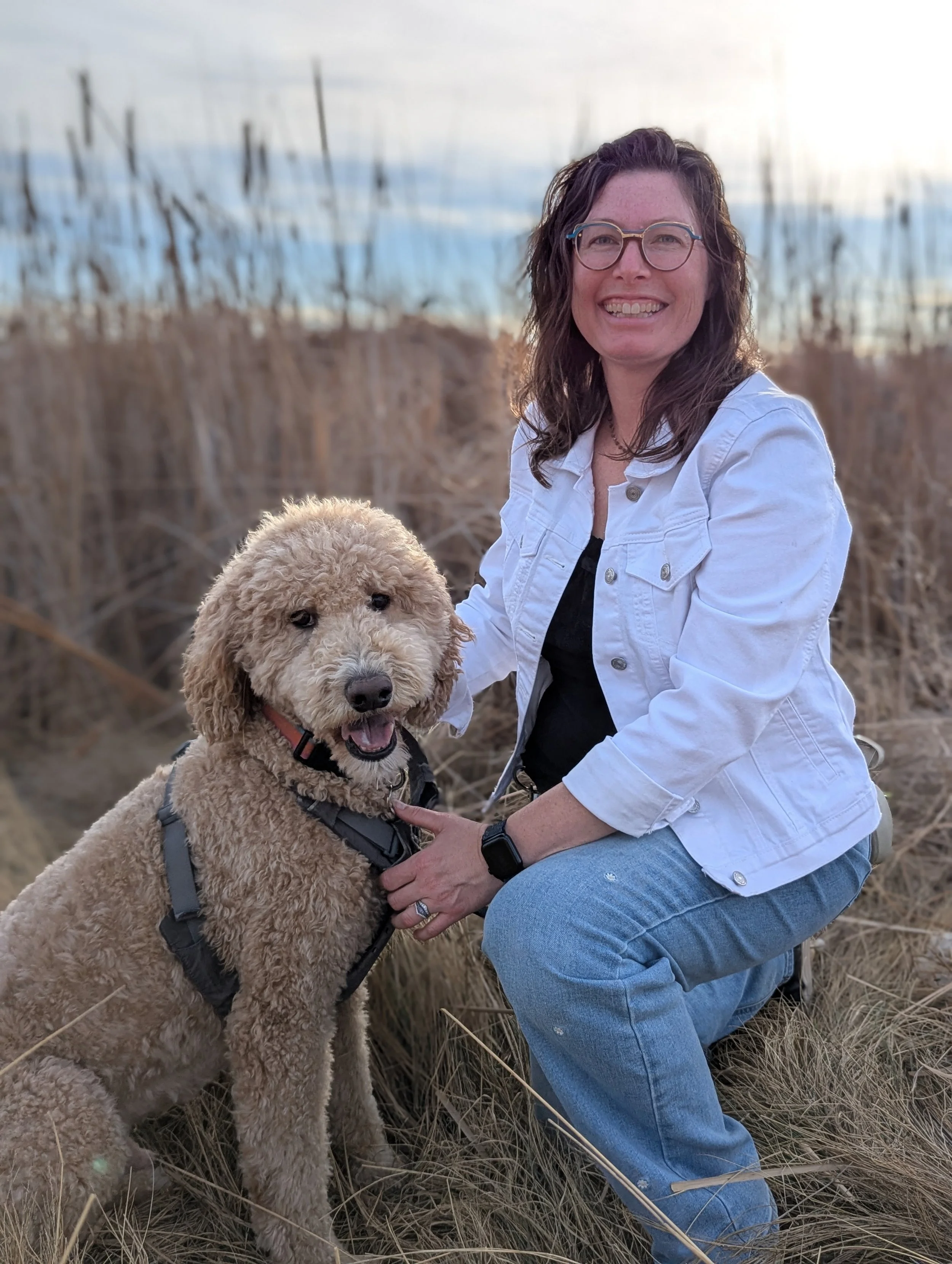 The author with glasses and wavy brown hair smiling while kneeling in a field of dry grass next to her large tan-colored goldendoodle dog during sunset.