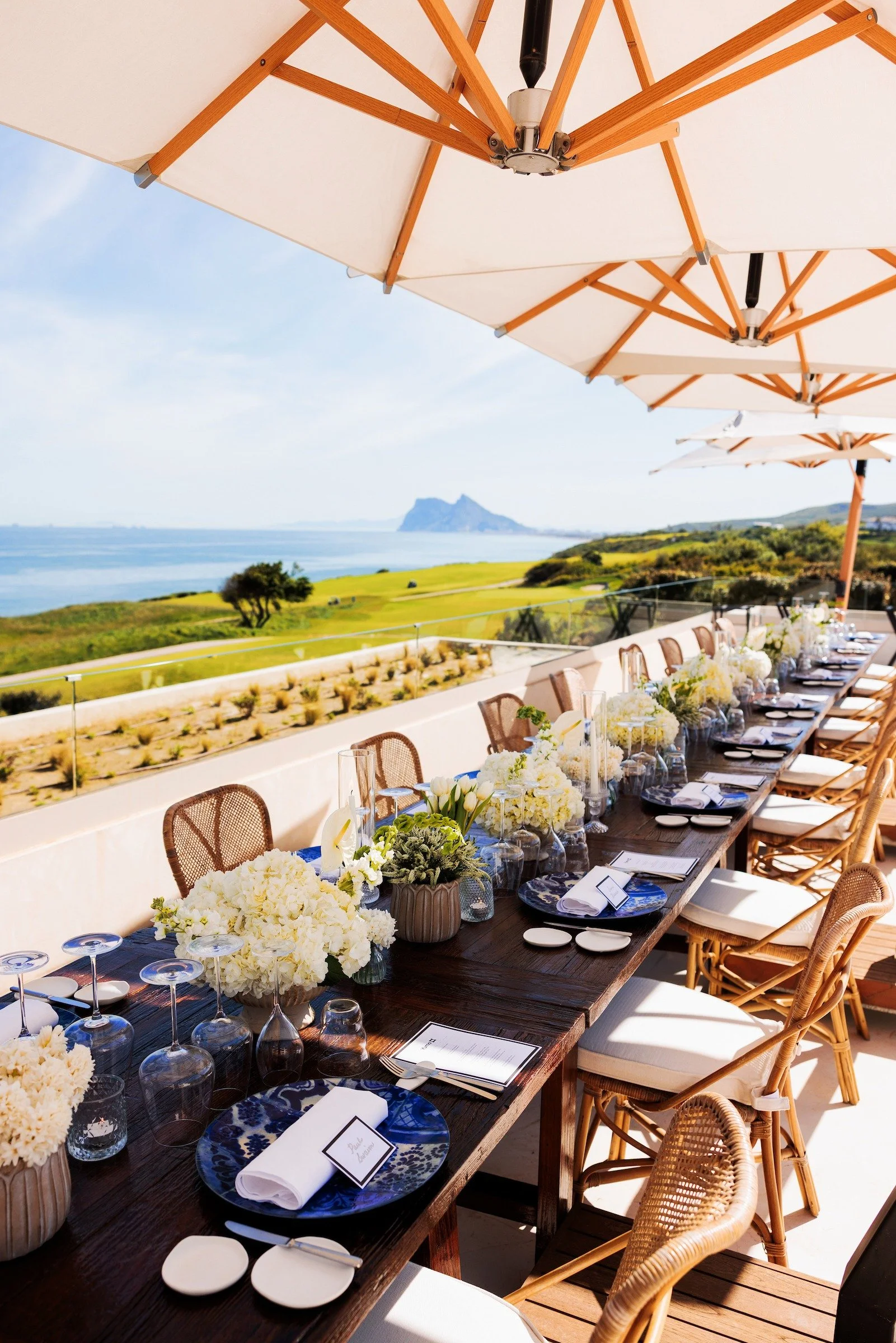 Luxury outdoor long table dinner setup with white floral centerpieces, blue place settings and panoramic Mediterranean views, corporate summit, Costa del Sol Spain — Grove Projects