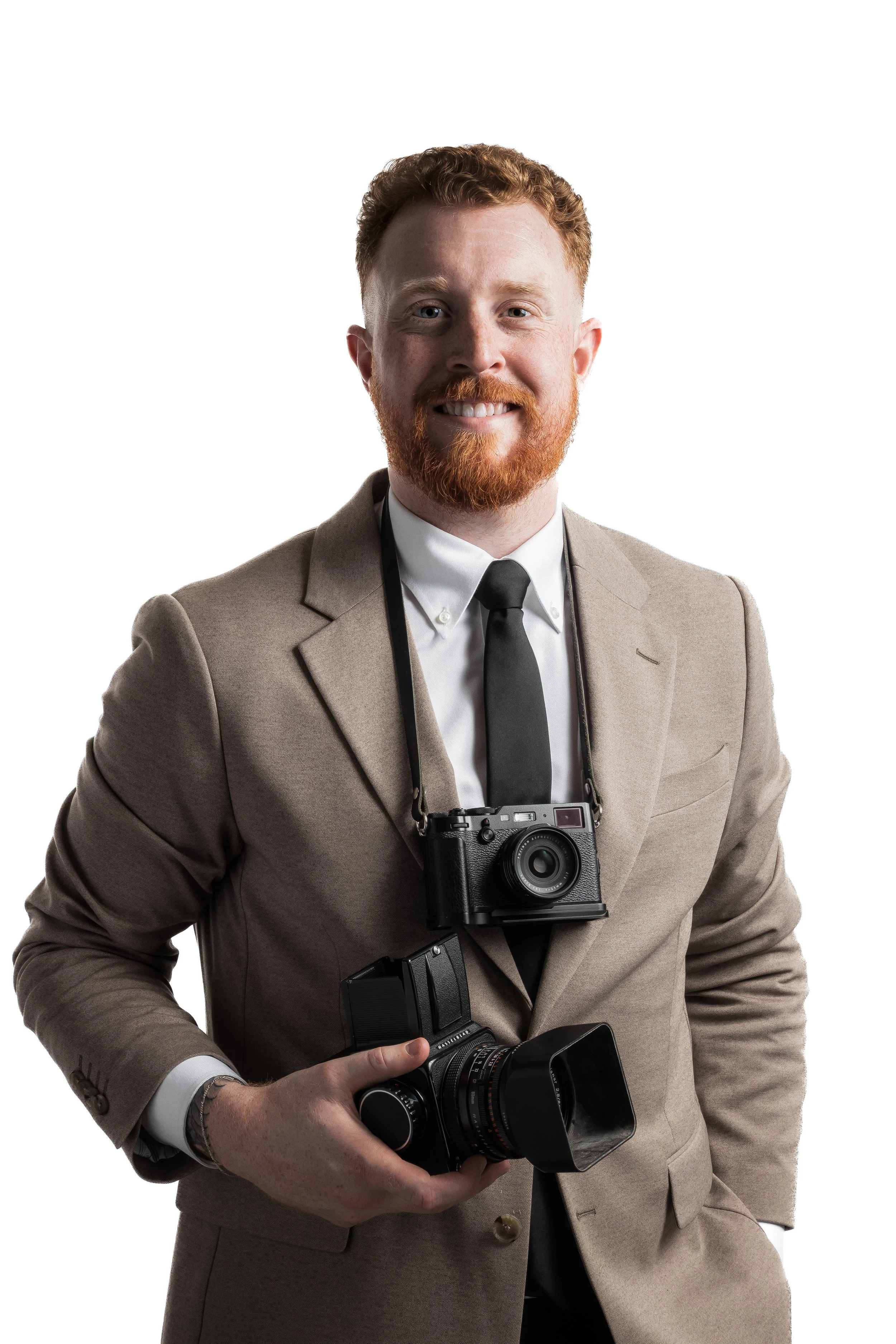 A man with red hair and a beard wearing a beige suit, holding a vintage camera in his hands, smiling at the camera, with a white background.