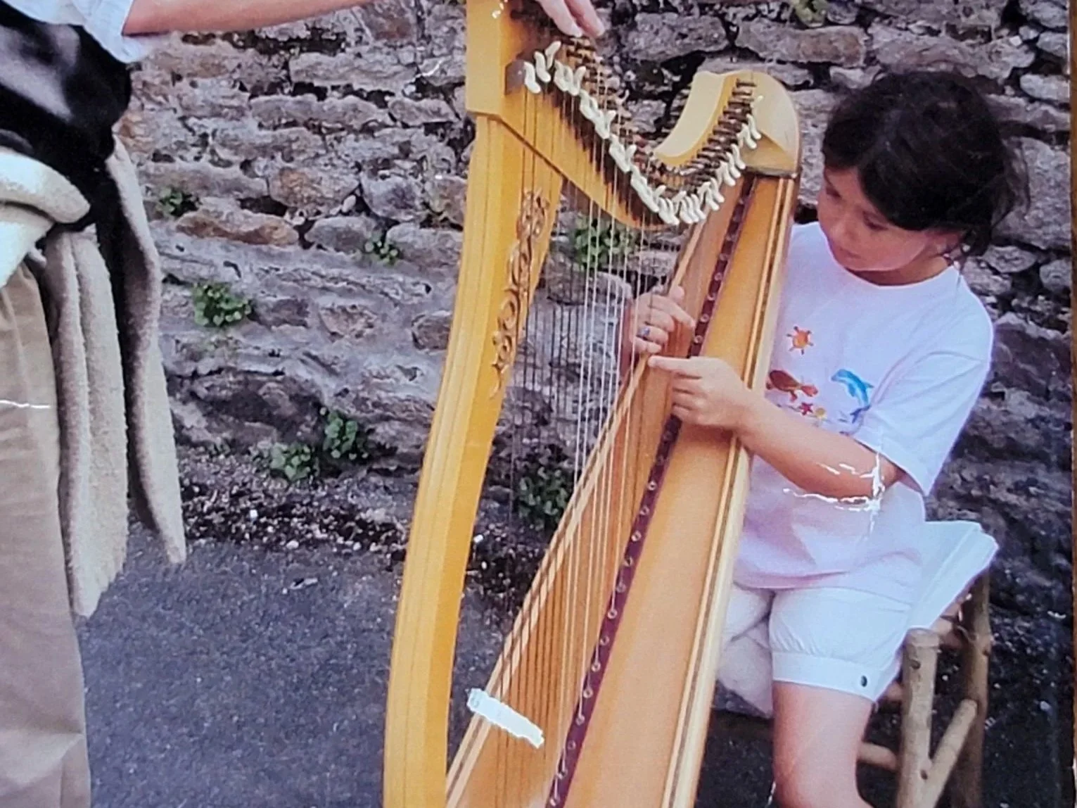 A young person playing a wooden harp outdoors near a stone wall.