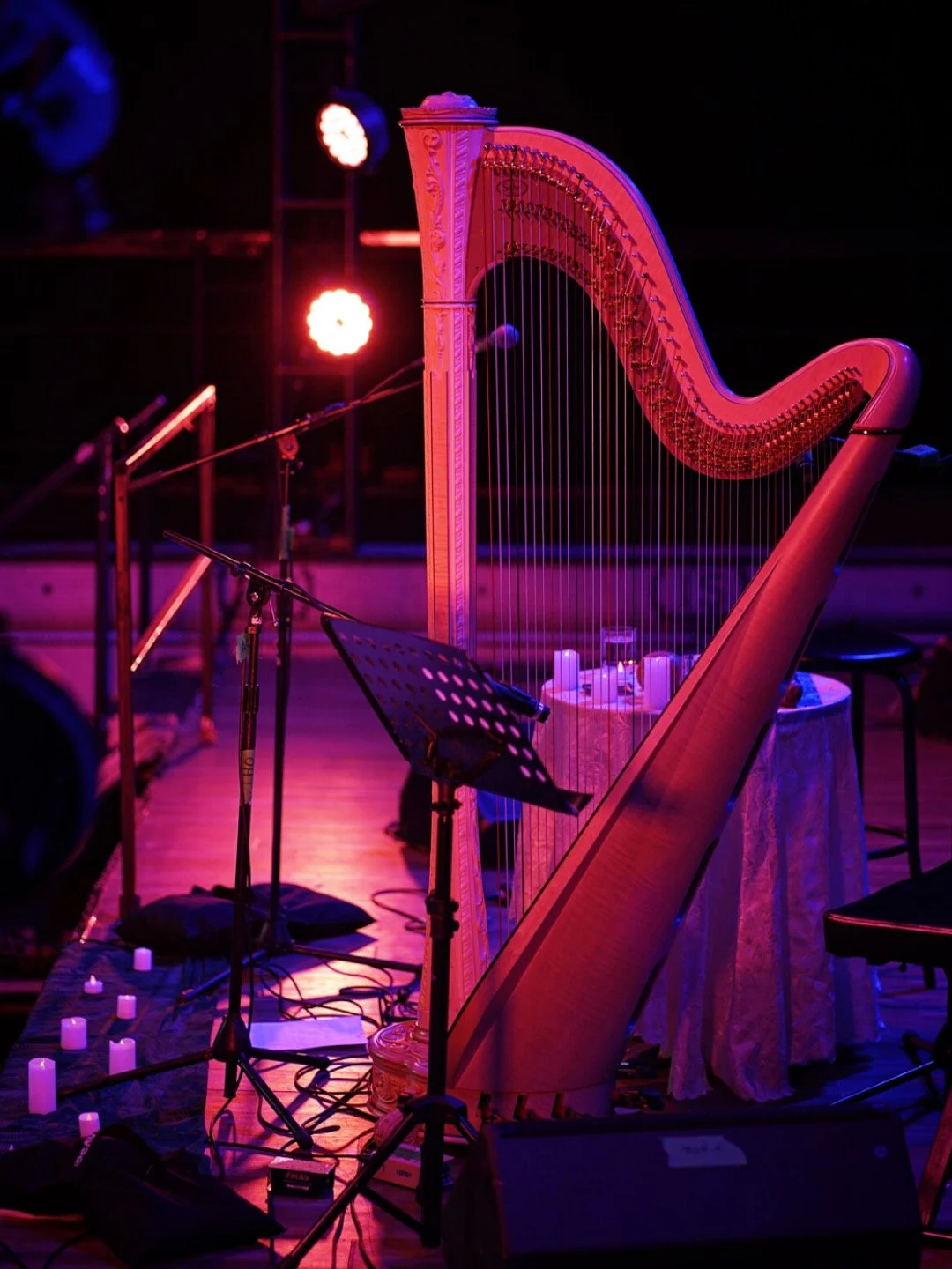 A harp on a stage illuminated with purple and pink lighting, surrounded by music stands, microphones, and small candle lights.
