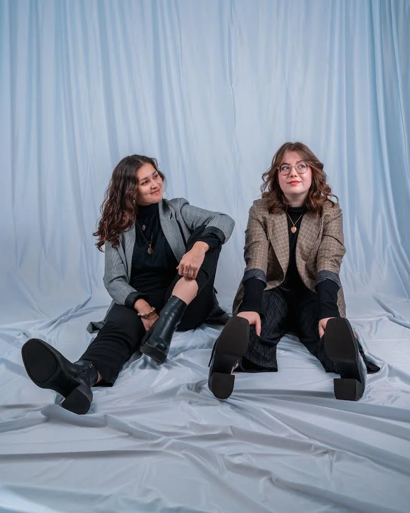 Two women sitting on a white cloth against a blue curtain background, dressed in casual office attire, looking in different directions.