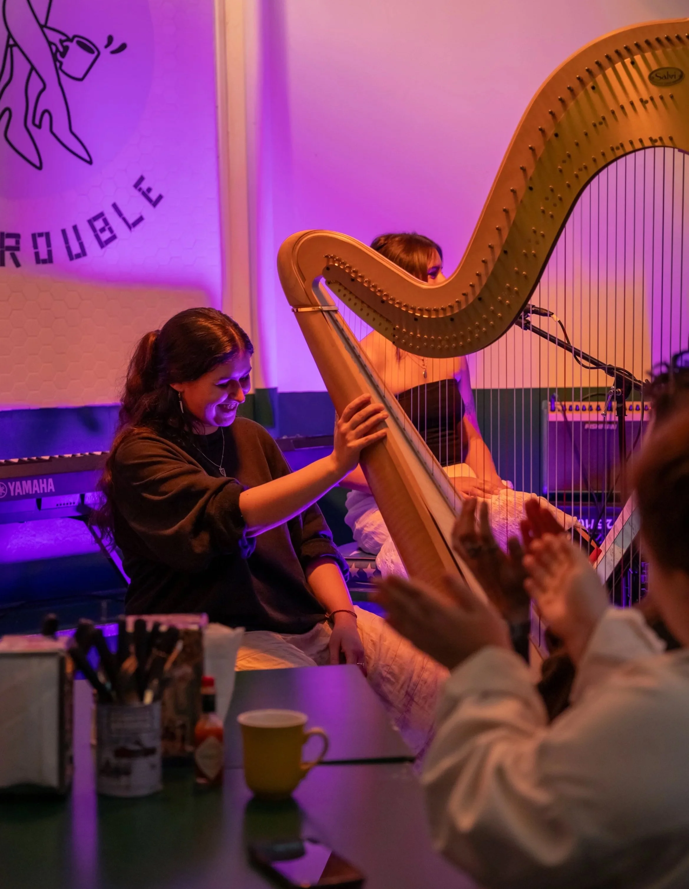 A woman playing a harp during a live performance in a room with purple lighting, with a woman in the background and various items on a table in the foreground.