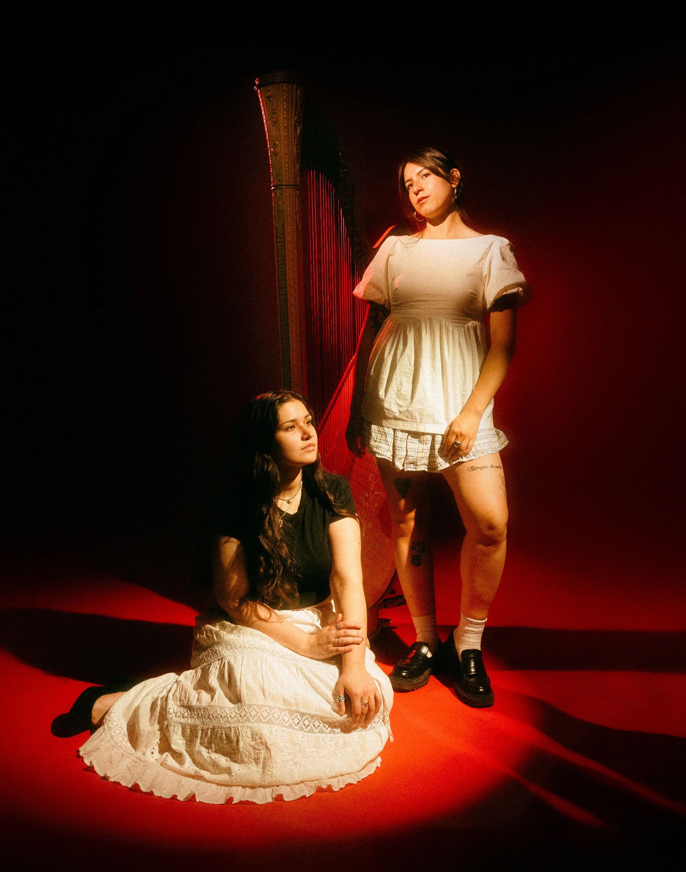 Two women posed in a dark room with red lighting. One woman is seated on the floor wearing a cream-colored dress, looking upwards, while the other woman stands behind her wearing a beige top with plaid shorts, looking sideways.