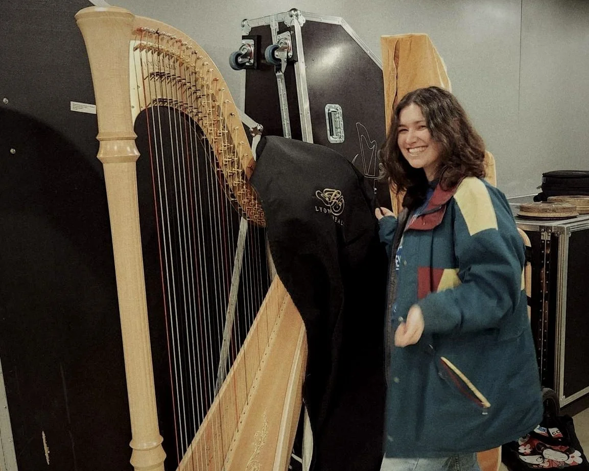 A young woman with curly hair smiling in front of a large, upright harp. She is wearing a colorful jacket, jeans, and black boots, and is standing in an indoor space, possibly a music or storage room.