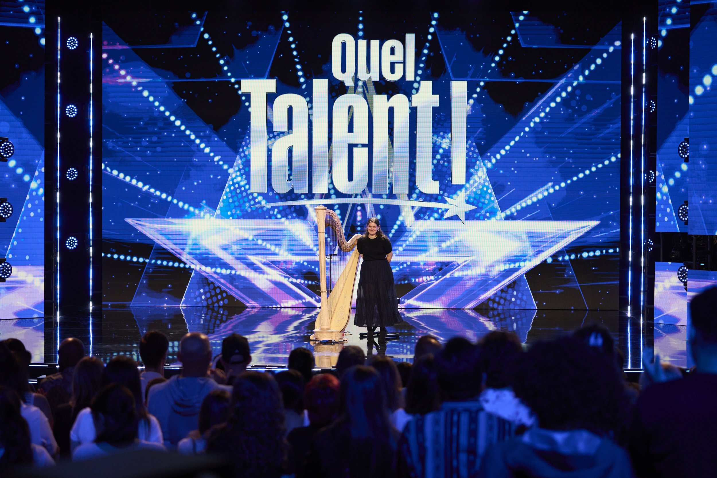 A woman standing on stage holding a harp at a talent show with the word 'QUEL TALENT' displayed on a large screen behind her, surrounded by blue lights and an audience watching.