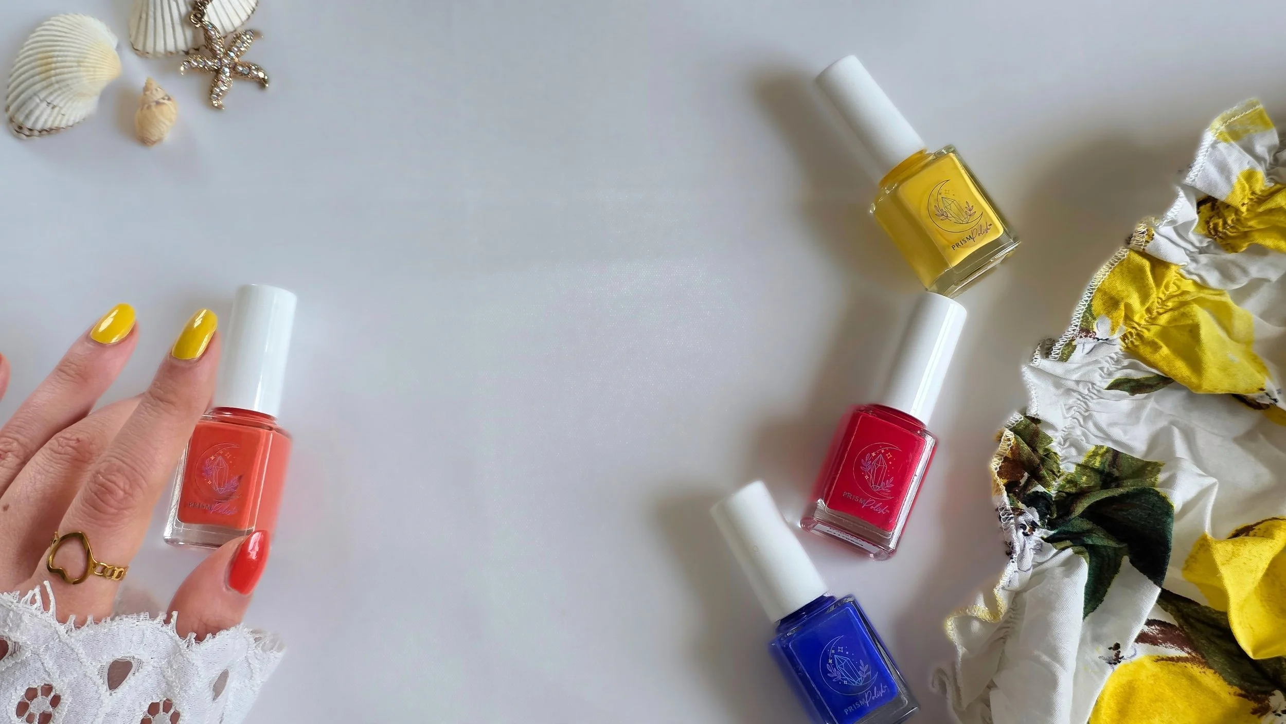 A person's hand with yellow and orange nail polish holding a red nail polish bottle. Several other nail polish bottles in yellow, red, and blue are scattered on a white surface. A yellow and floral cloth is partially visible on the right side. In the top left corner, there are seashells and a starfish.