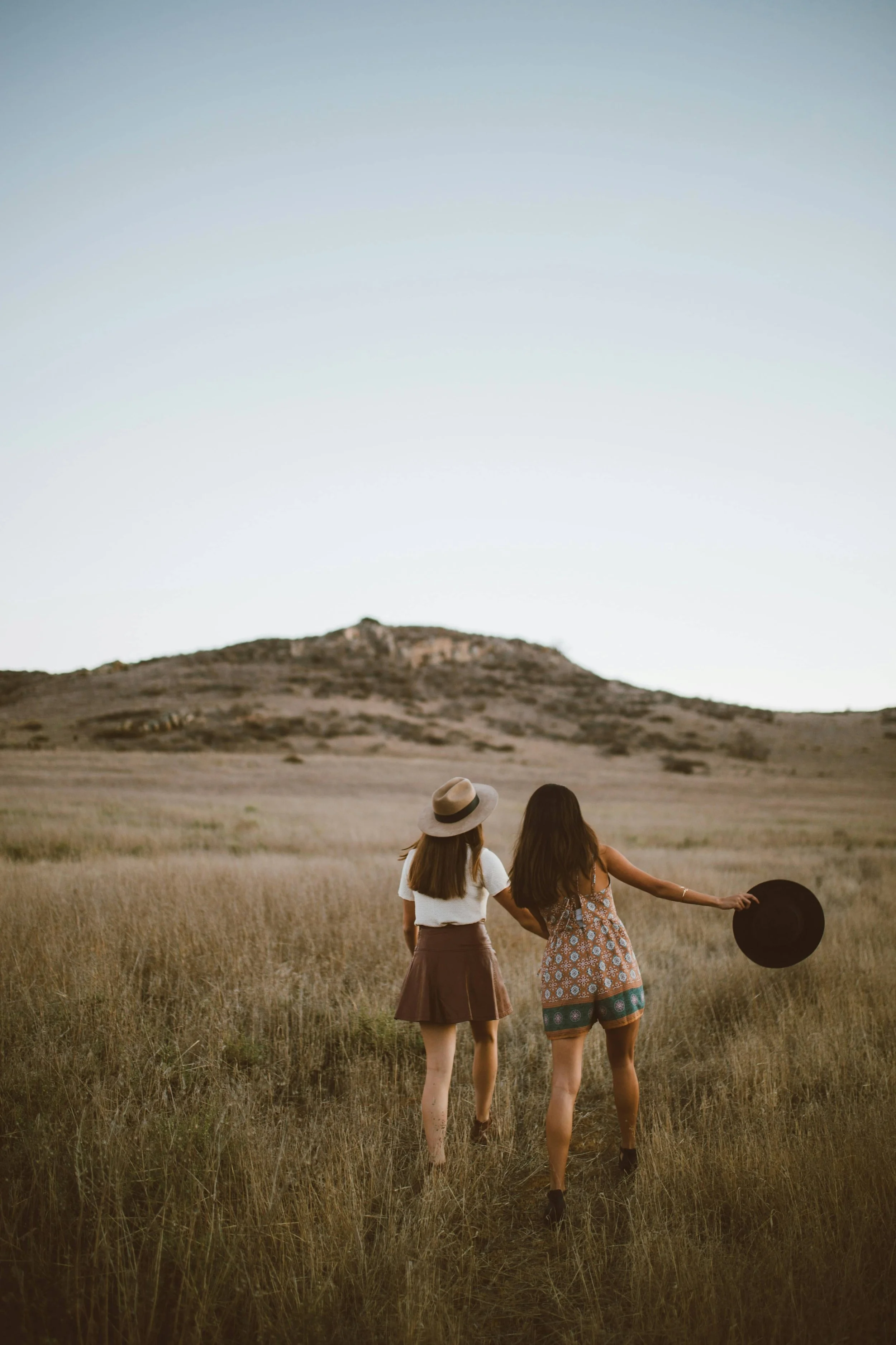 Two young women walk hand-in-hand through a grassy field towards a hill, one wearing a hat and the other holding a hat in her hand, during sunset or sunrise.