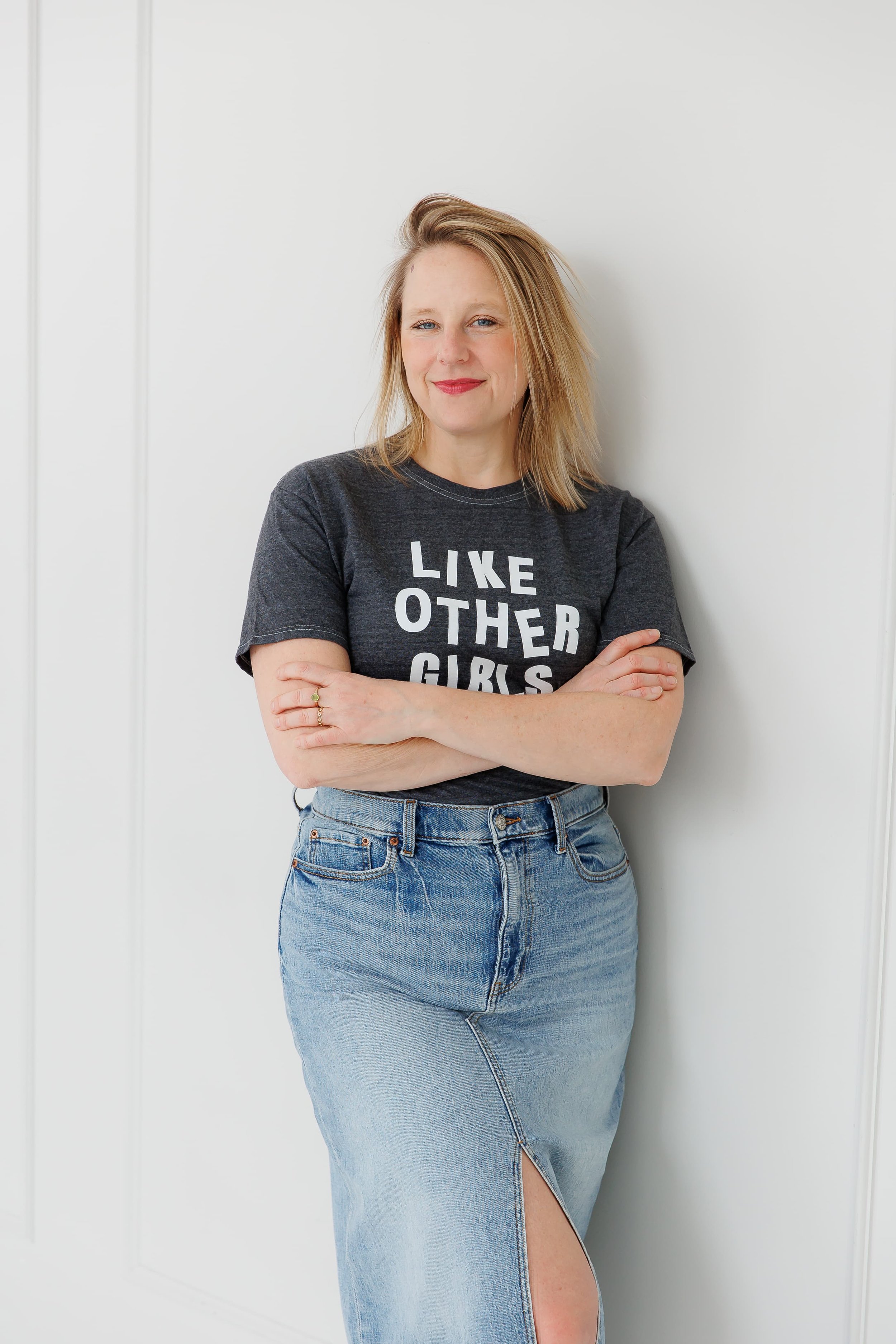 Andrea Tessier, a woman with blonde hair, wearing a dark gray T-shirt with the text 'LIKE OTHER GIRLS', and a high slit on her jean skirt, standing with crossed arms against a plain white wall, smiling.