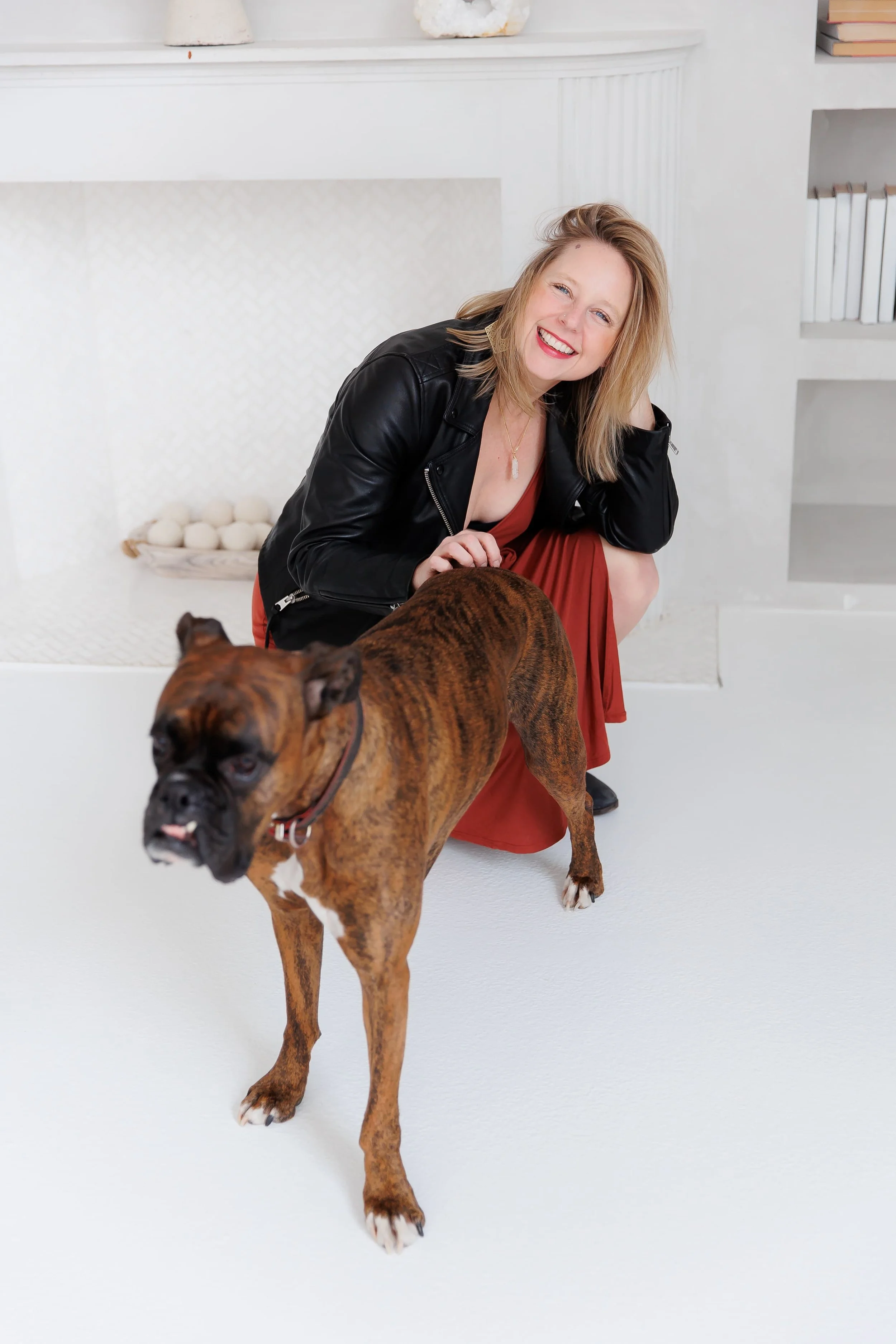 Andrea Tessier, IFS coach, a woman with blonde hair wearing a black leather jacket and a red dress, smiling and squatting in front of a brown boxer dog, standing on a white floor with white background.