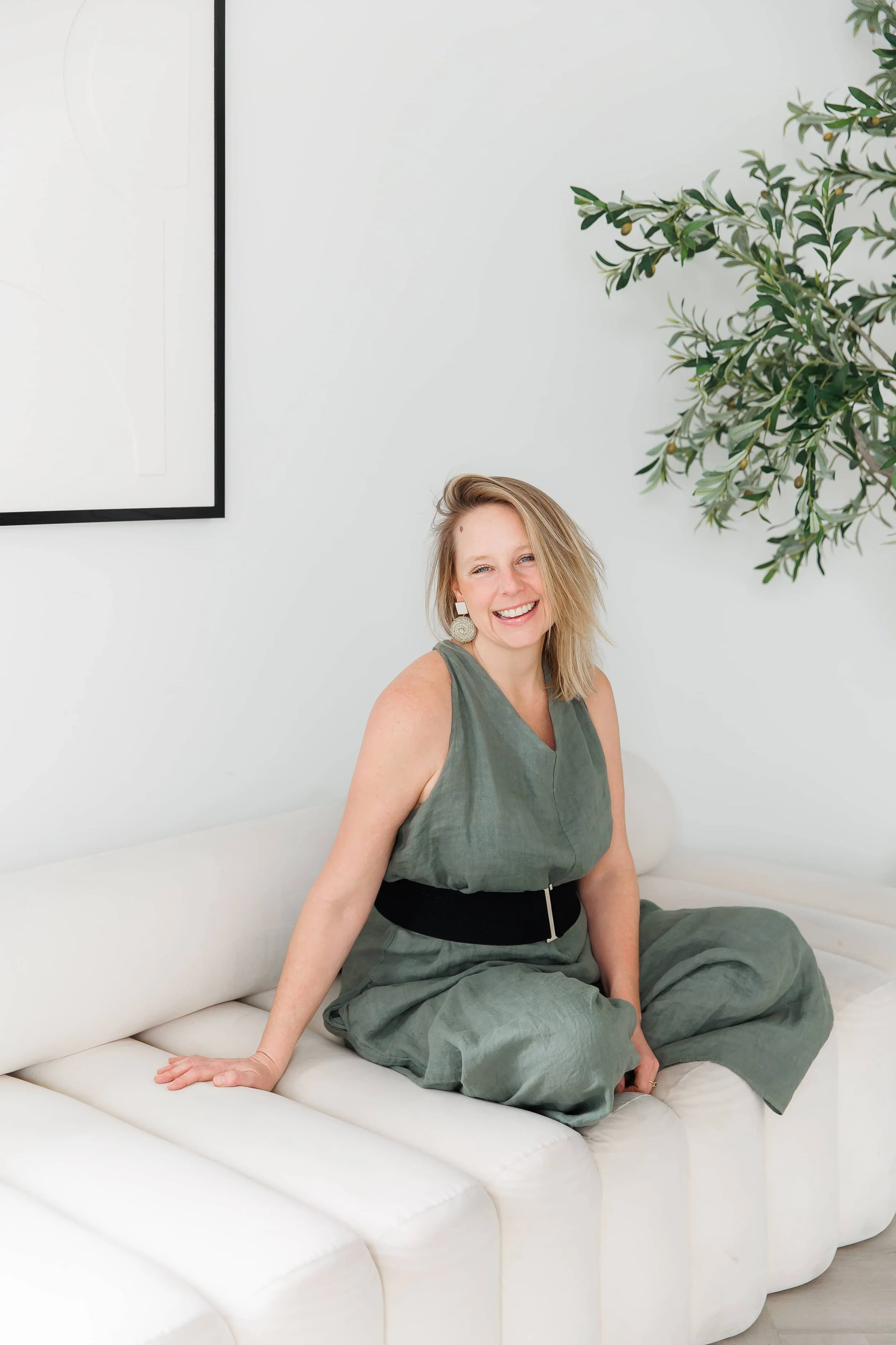 Andrea Tessier, a shadow work coach, with blonde hair and earrings sitting on a white sofa in a room with white walls, a large framed picture, and a green leafy plant.
