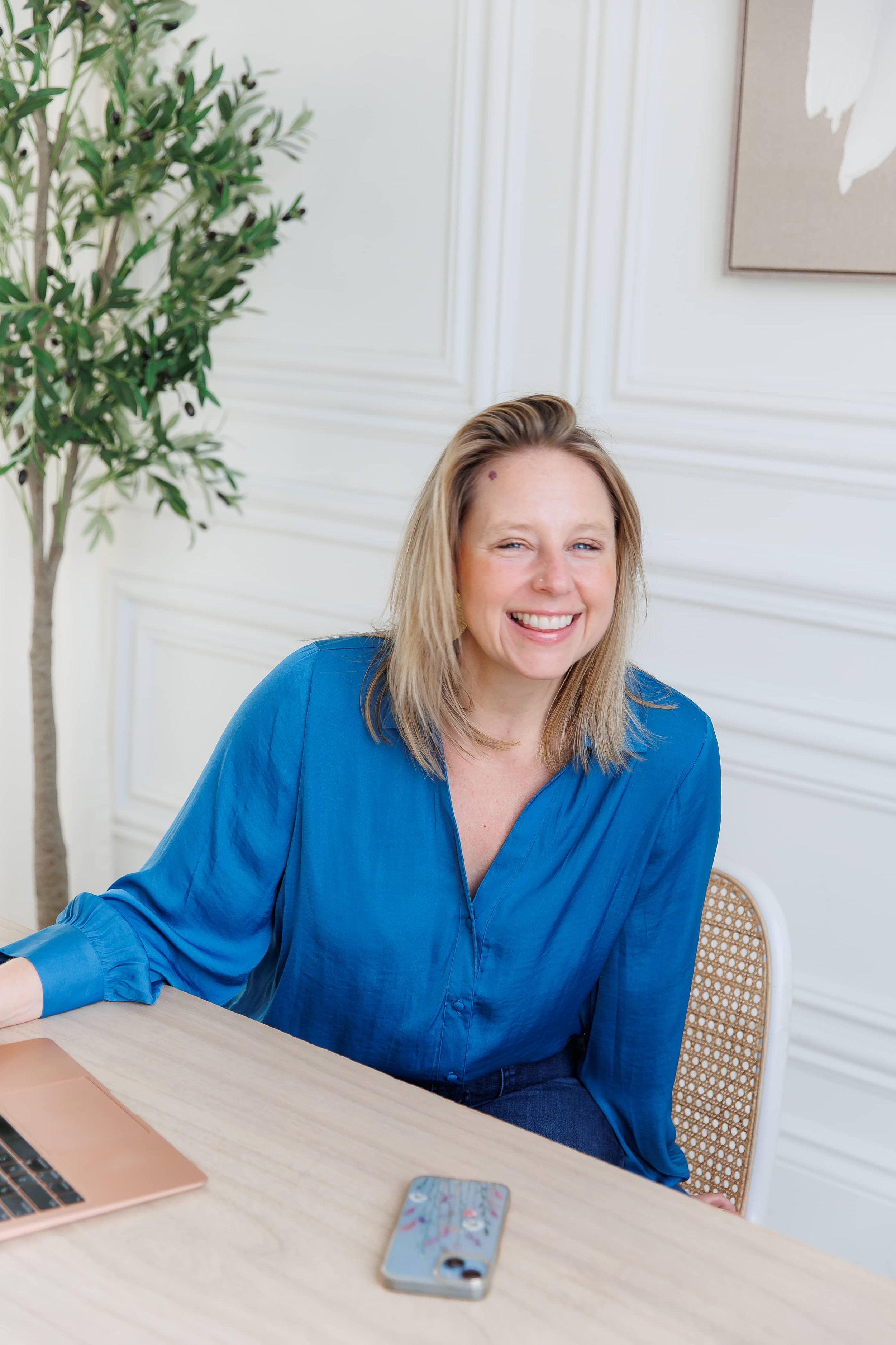 Andrea Tessier, a coach for high achieving women, with blonde hair and a blue blouse sitting at a light wood table, smiling, with a smartphone and a rose gold laptop on the table. There is a plant and framed art on a white wall behind her.