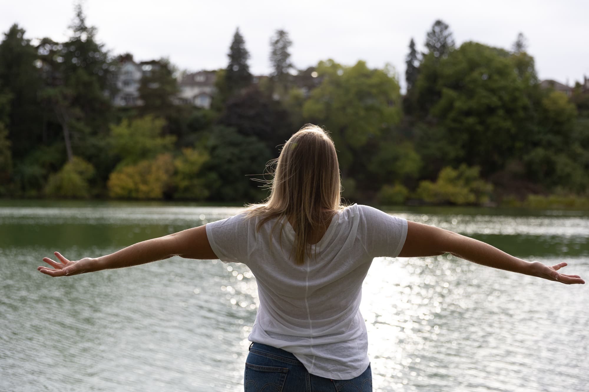 Andrea Tessier, a spiritual coach, based in Toronto, with long hair standing by a lake with arms outstretched, facing the water and trees on the opposite shore.
