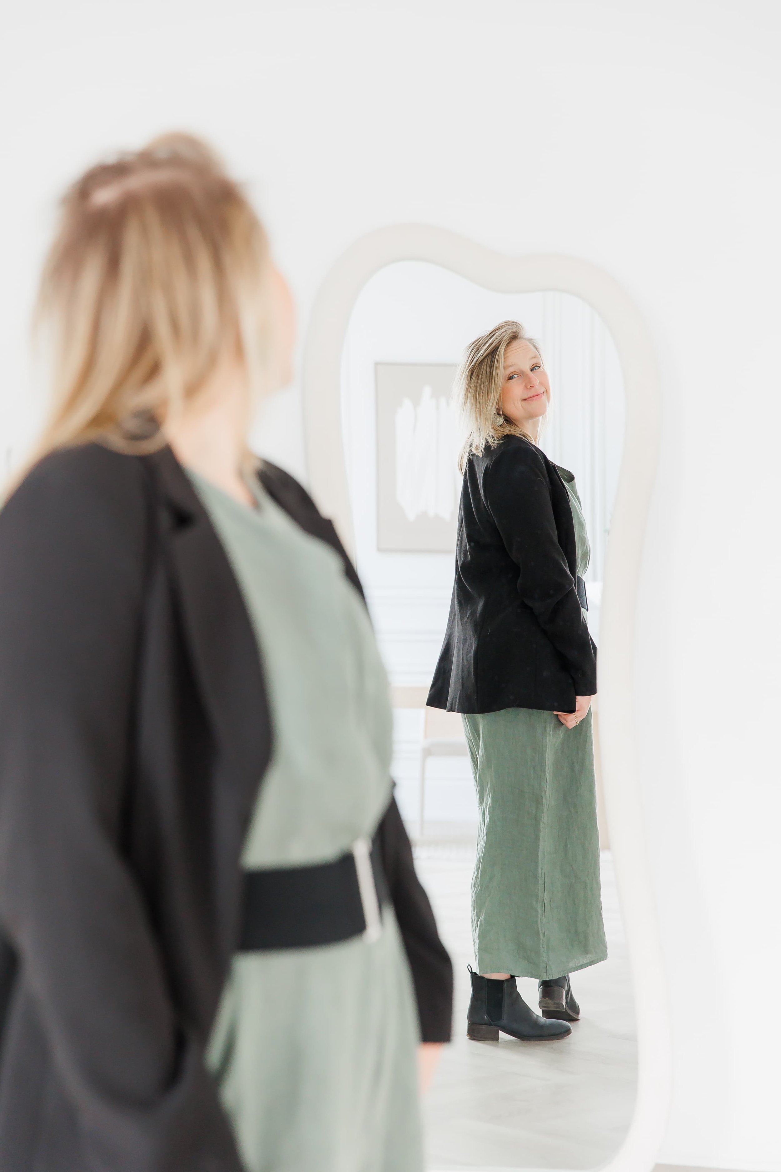 Andrea Tessier, a woman with blonde hair and blue eyes is standing and looking into a mirror, wearing a black blazer and a green dress. The reflection shows her smiling slightly, wearing black boots, in a bright, white room with minimalistic decor.