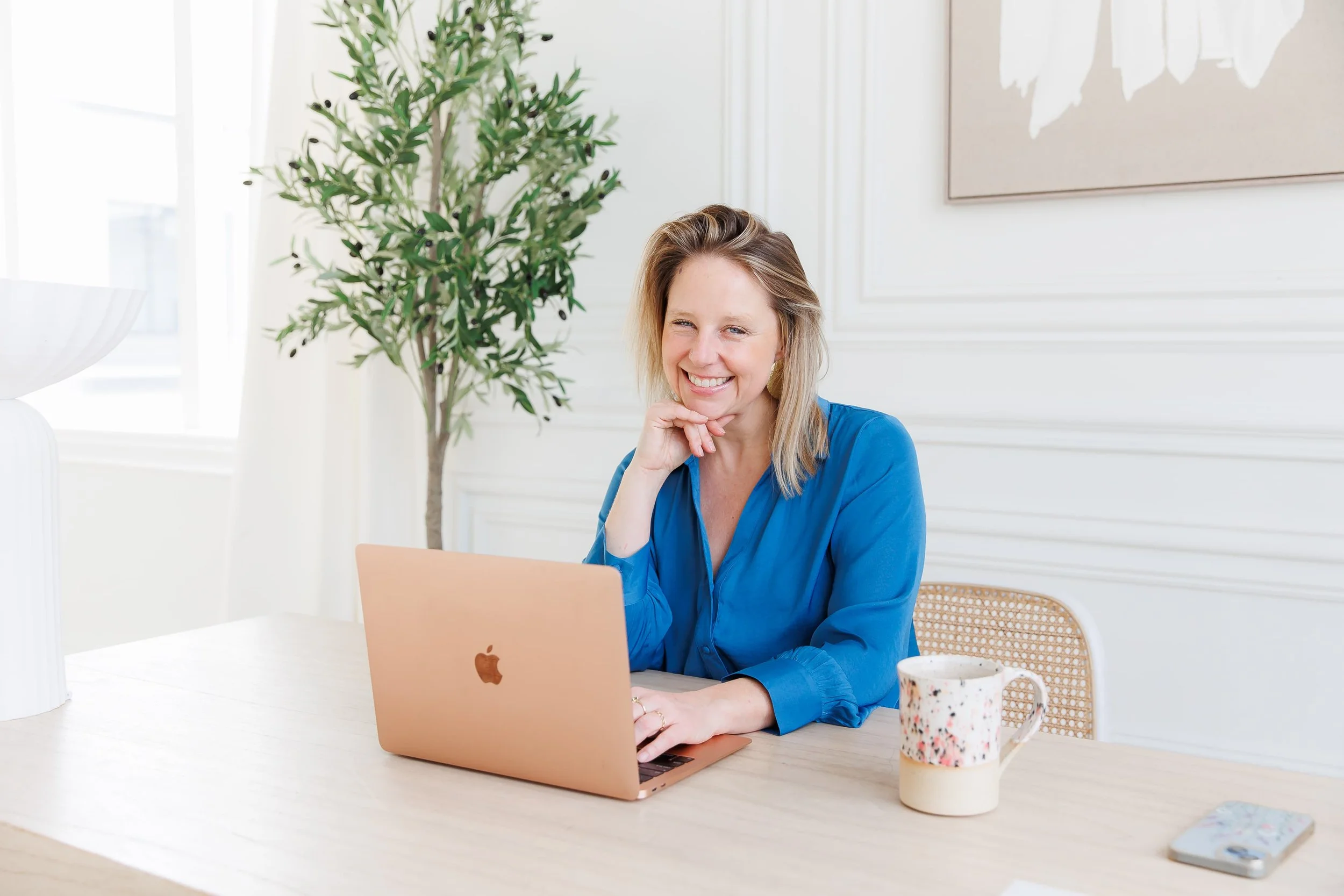 Andrea Tessier, a leadership coach, smiling and sitting at a table with a rose gold MacBook. There is a mug with a speckled design, a smartphone, and a plant in the background. The room has white walls and a window letting in natural light.