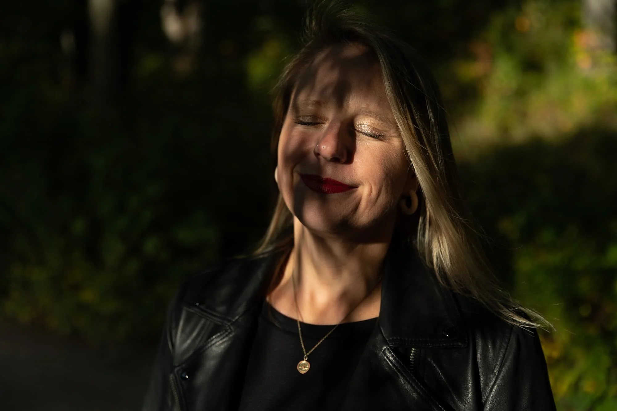 Andrea Tessier, a high capacity coach for women, with blonde hair and red lipstick smiling with eyes closed, wearing a black leather jacket and a gold necklace, outdoors with blurred green foliage in the background.