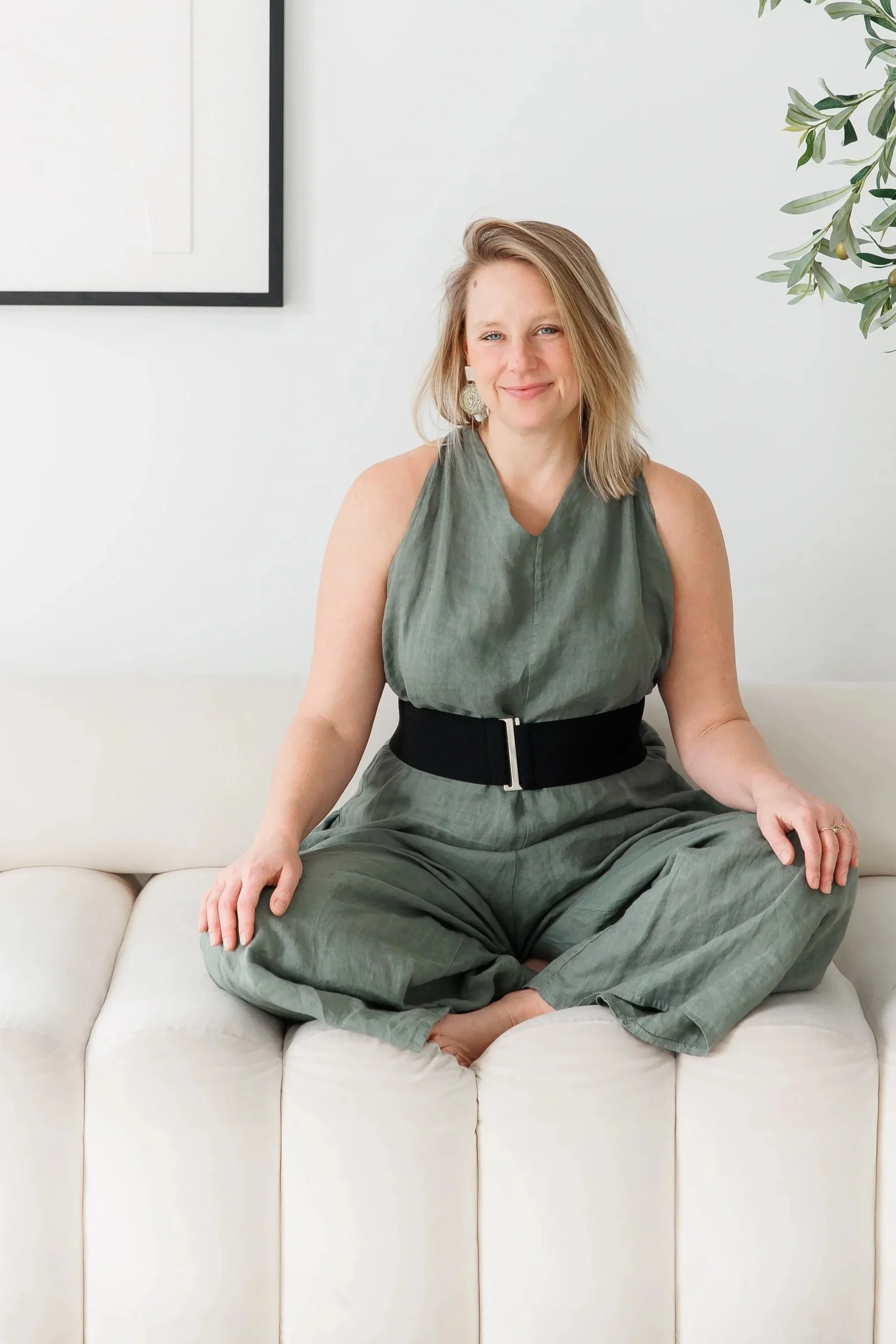 Andrea Tessier, high capacity coach. Sitting cross-legged on a cream-colored sofa, smiling at the camera, with a white wall and some greenery in the background.