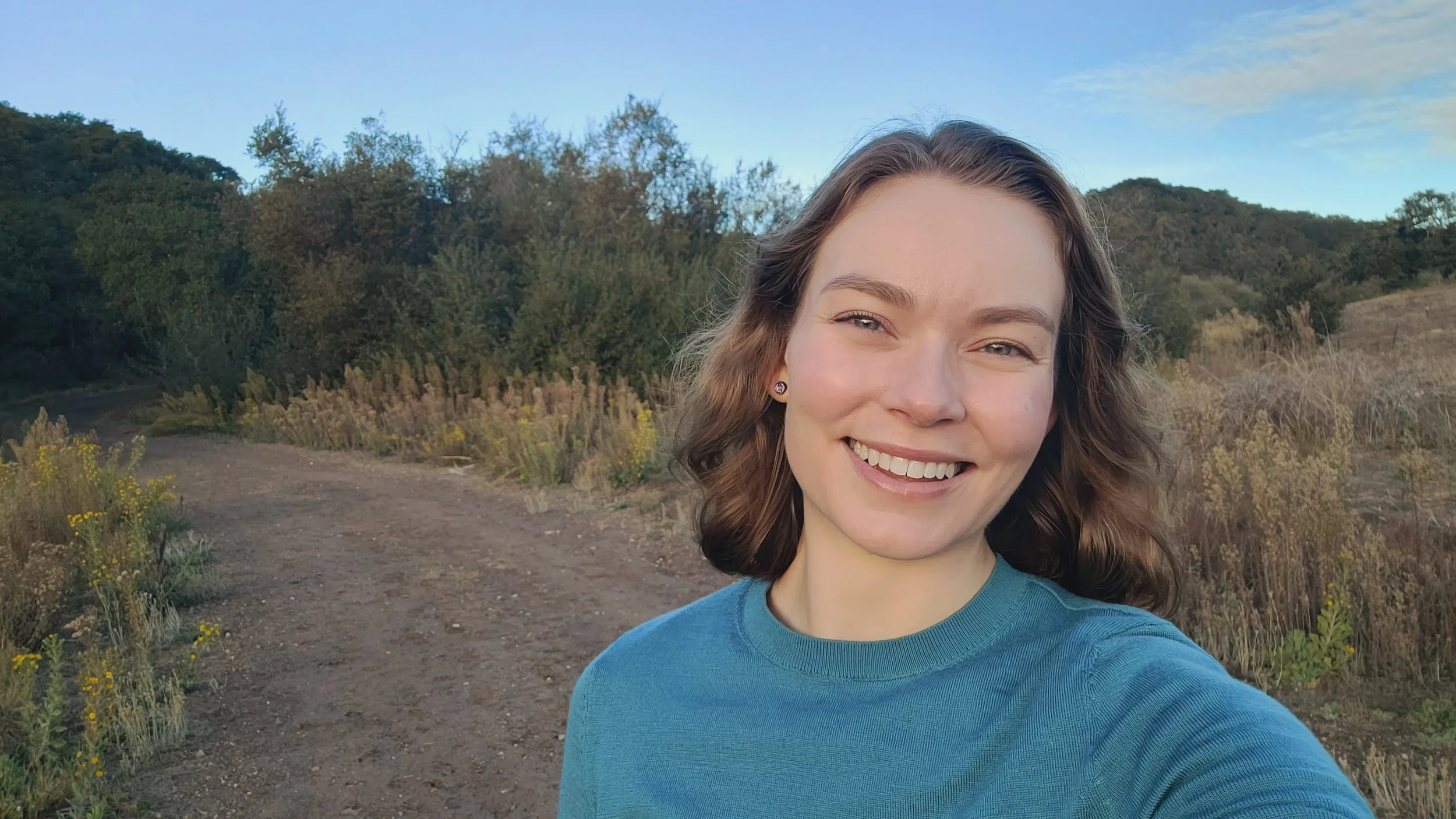 A client of Andrea Tessier, leadership coach. A smiling young woman with curly brown hair and earrings taking a selfie outdoors on a trail, with green hills, trees, and a cloudy sky in the background.