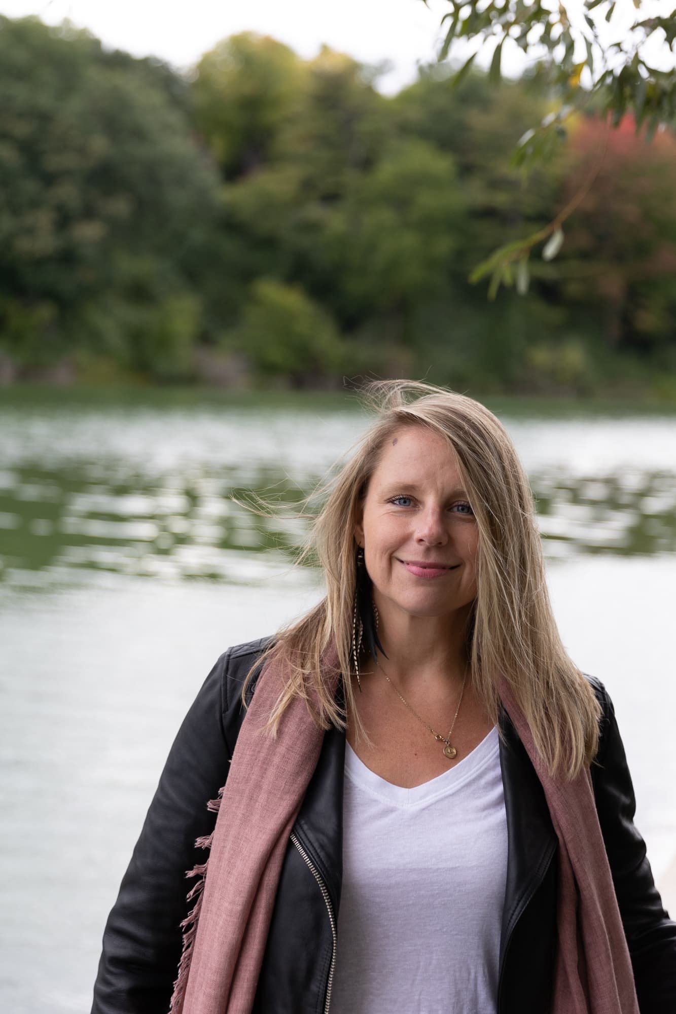 Andrea Tessier, women's coach, with long blonde hair is standing outdoors near a body of water with trees in the background. She is smiling, wearing a black jacket, white top, and a pink scarf.