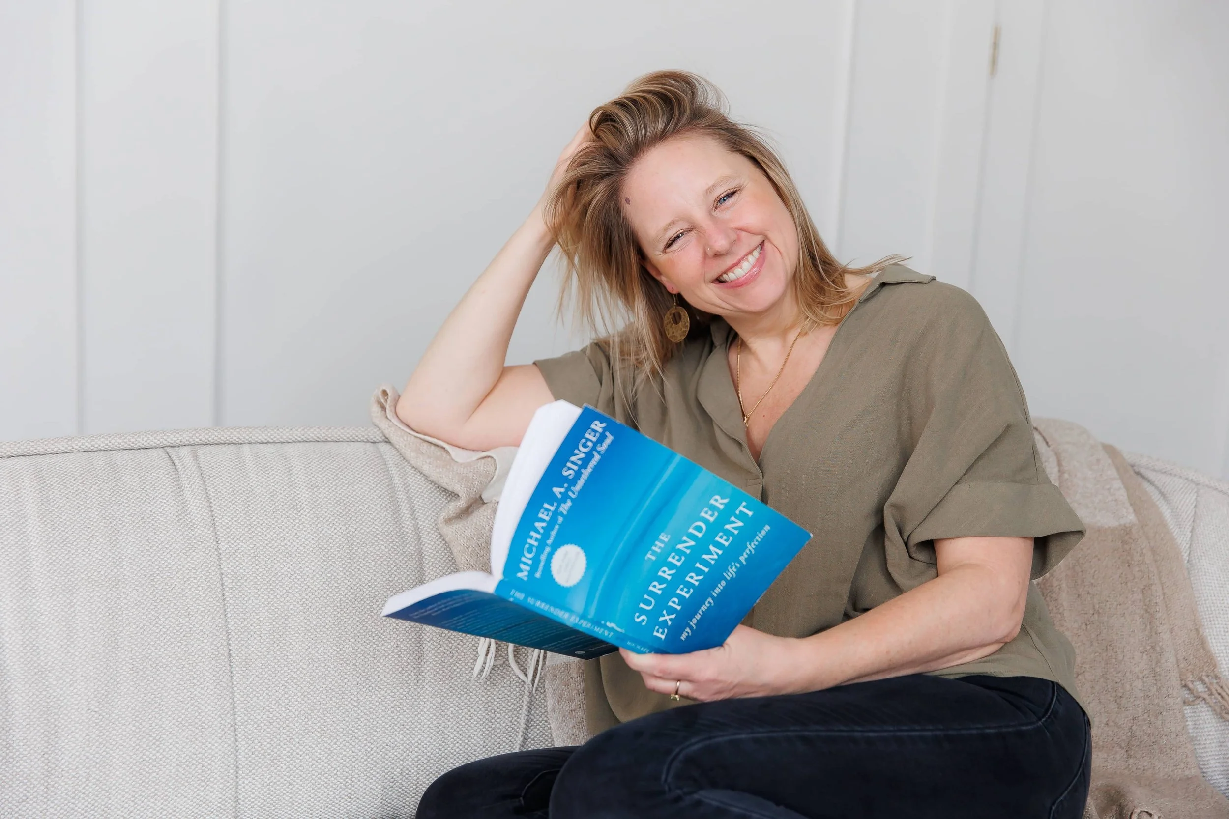 Andrea Tessier, a Toronto-based life coach, with blonde hair and earrings sitting on a beige couch, smiling and looking at the camera, holding a blue book titled 'The Confidence Solution' by Michael A. Singer.