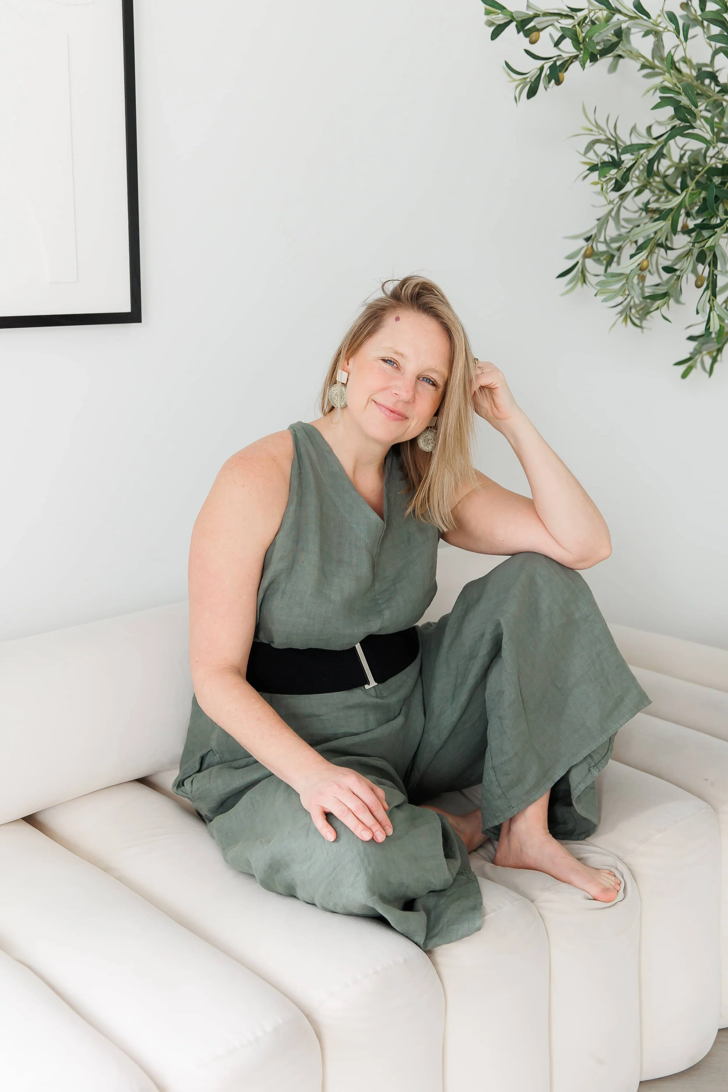 Andrea Tessier, somatic coach for women, with blonde hair wearing a green dress, sitting on a white leather sofa, smiling and looking at the camera, with one arm resting behind her head and the other on her knee.