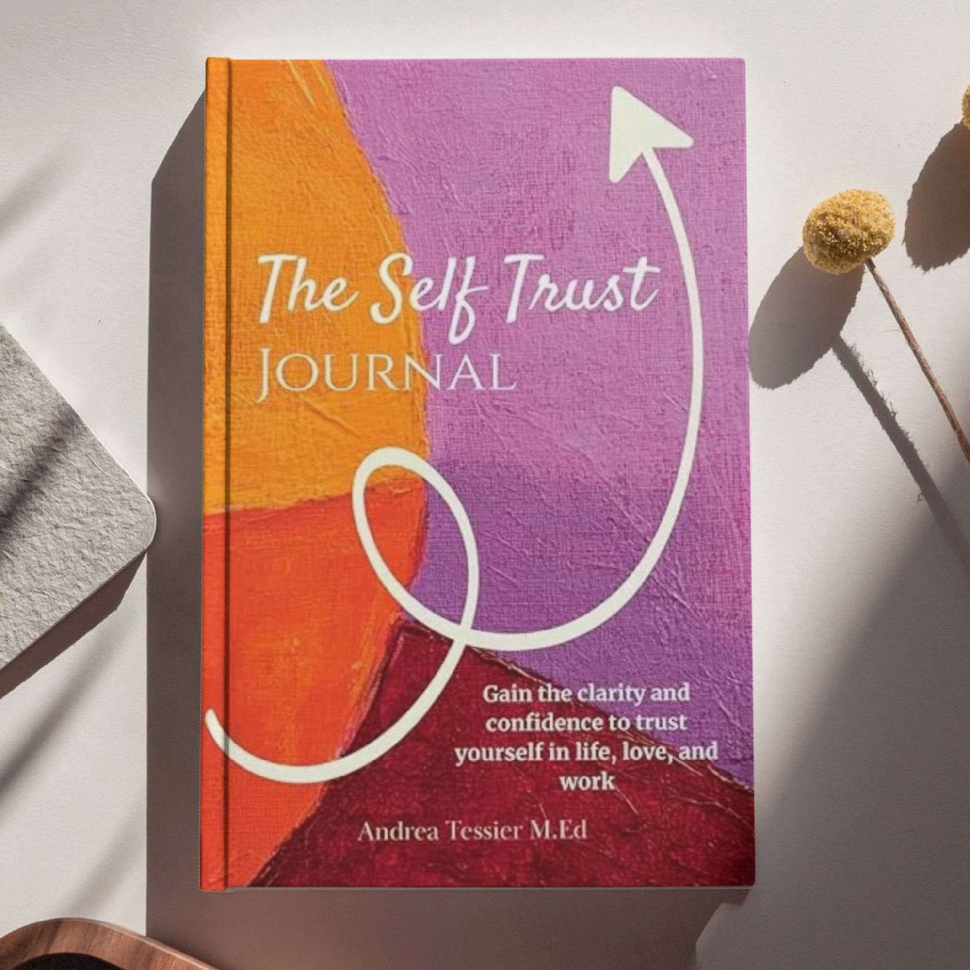 Close-up of a colorful book cover titled 'The Self Trust Journal' with a graphic of a white arrow on a colorful background. The lower part of the cover shows a subtitle encouraging clarity and confidence in oneself, authored by Andrea Tessier M.Ed.