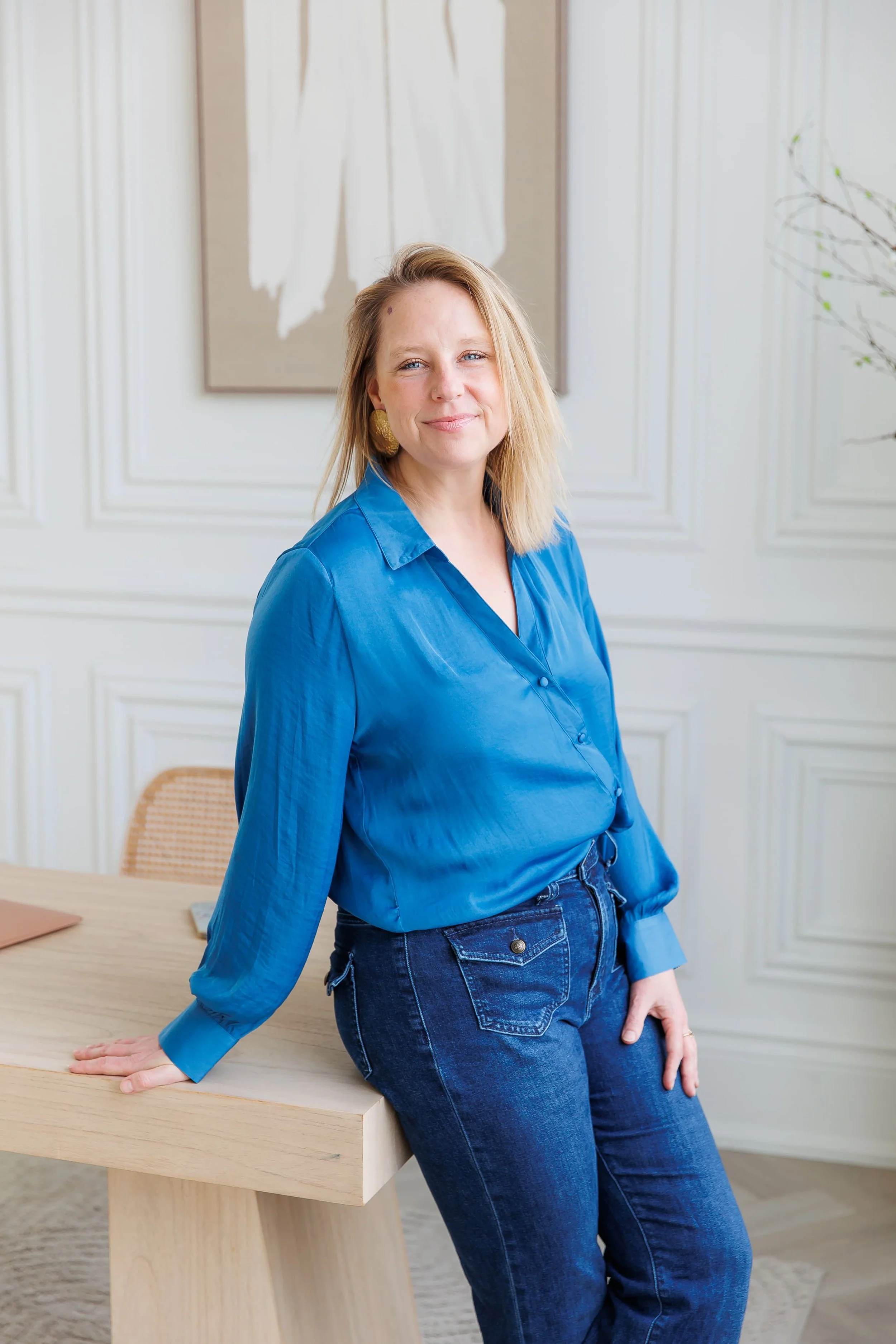 Andrea Tessier, a woman with blonde hair and blue eyes wearing a blue button-up shirt and dark jeans, leaning on a light-colored wooden table in a room with white paneled walls and framed artwork.