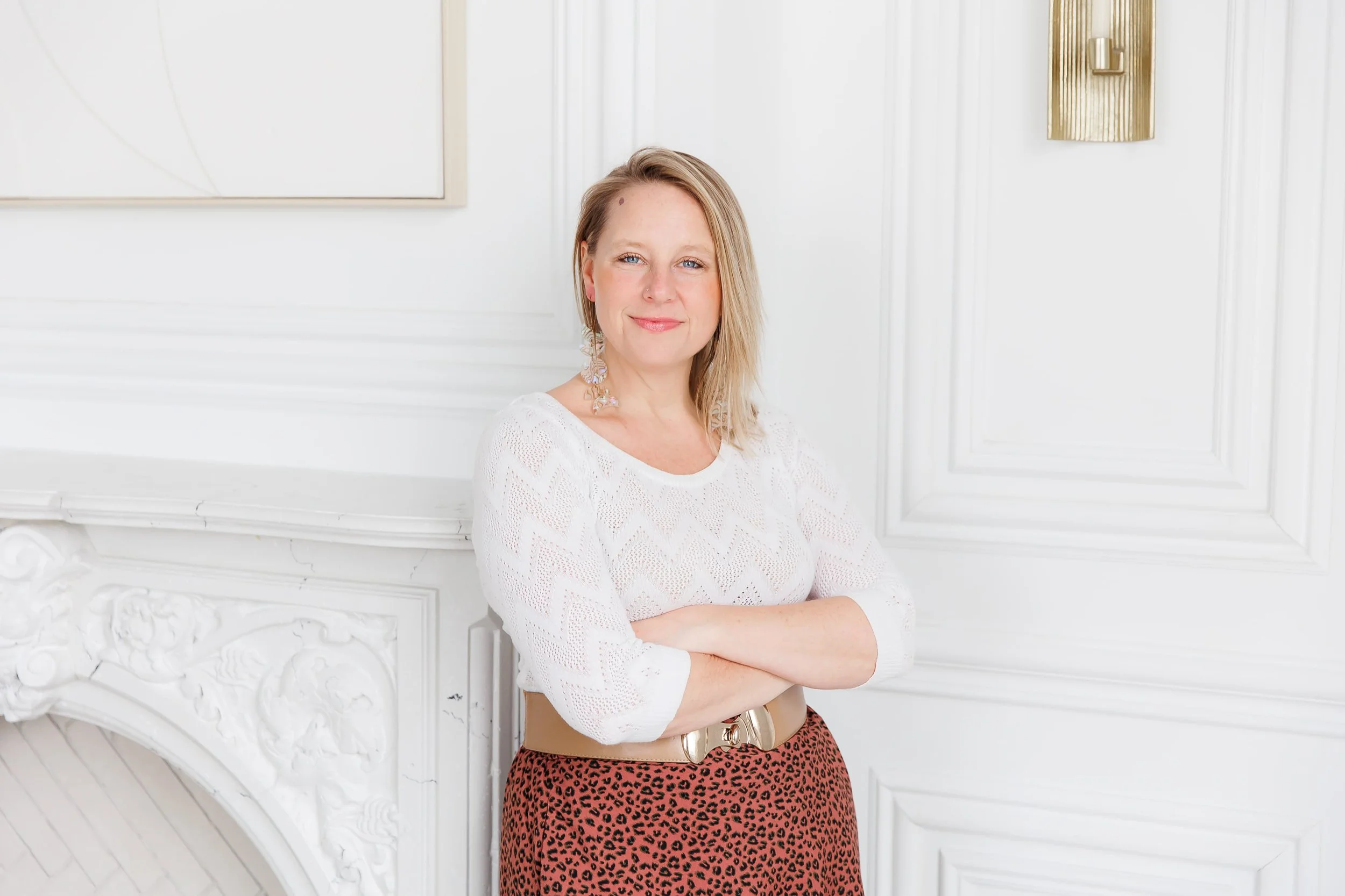 Andrea, a life coach for women, with blonde hair and blue eyes indoors, standing in front of ornate white paneling and a decorative wall sconce, wearing a white blouse with lace details and leopard print skirt.