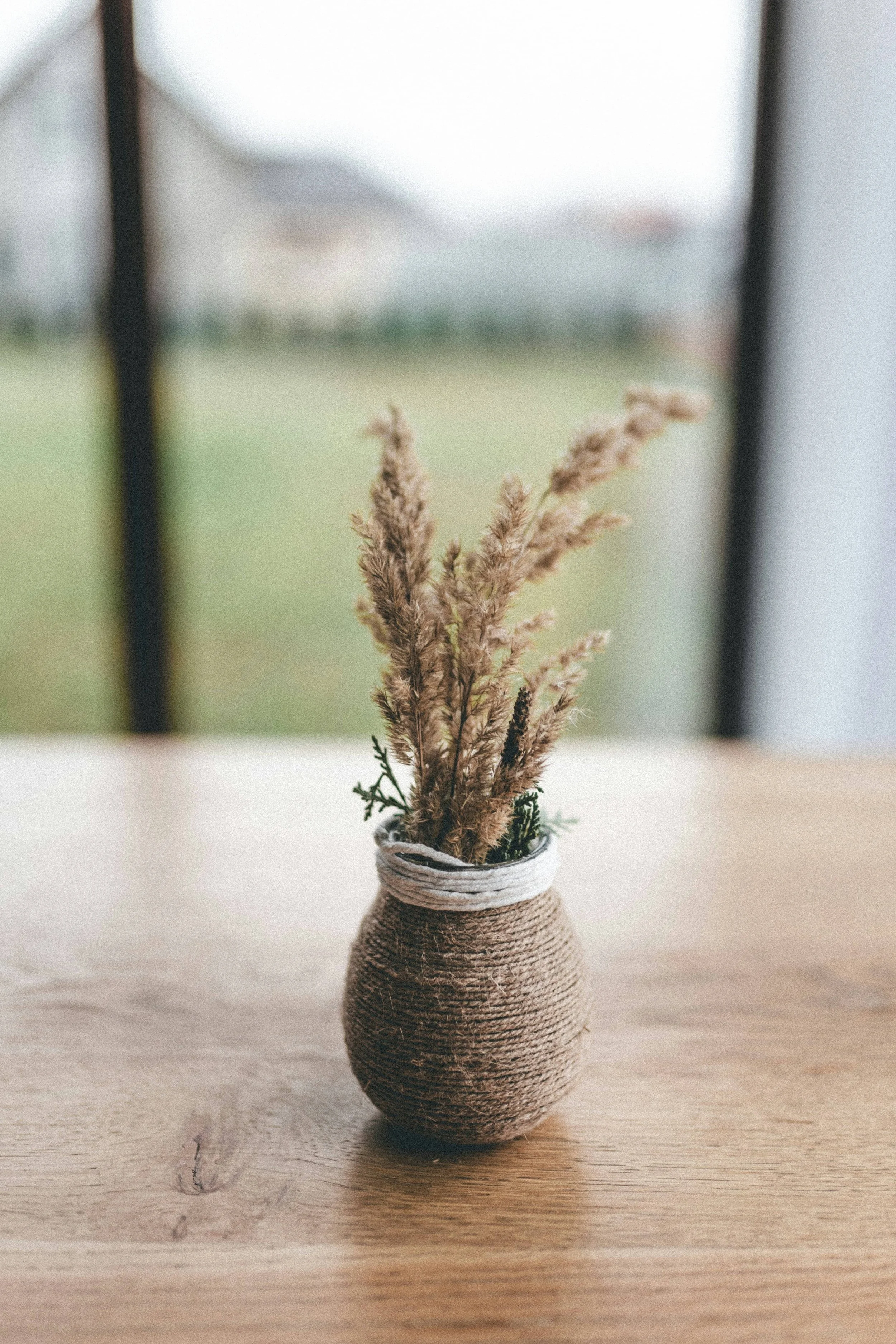 A small, round vase wrapped in twine with dried grasses and small greenery inside, placed on a wooden surface.